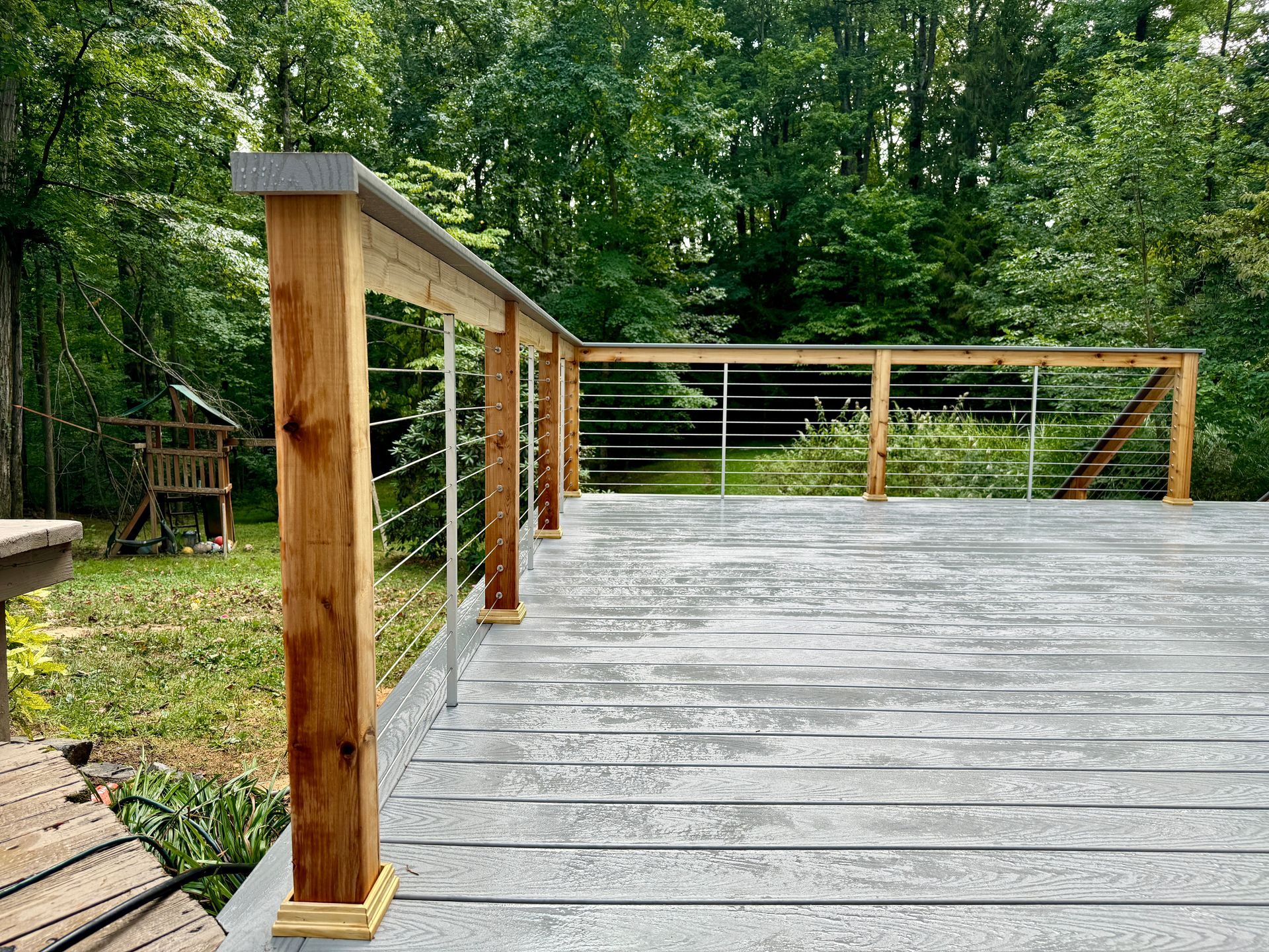 Deck with wooden posts and cable railings, overlooking a backyard with trees.