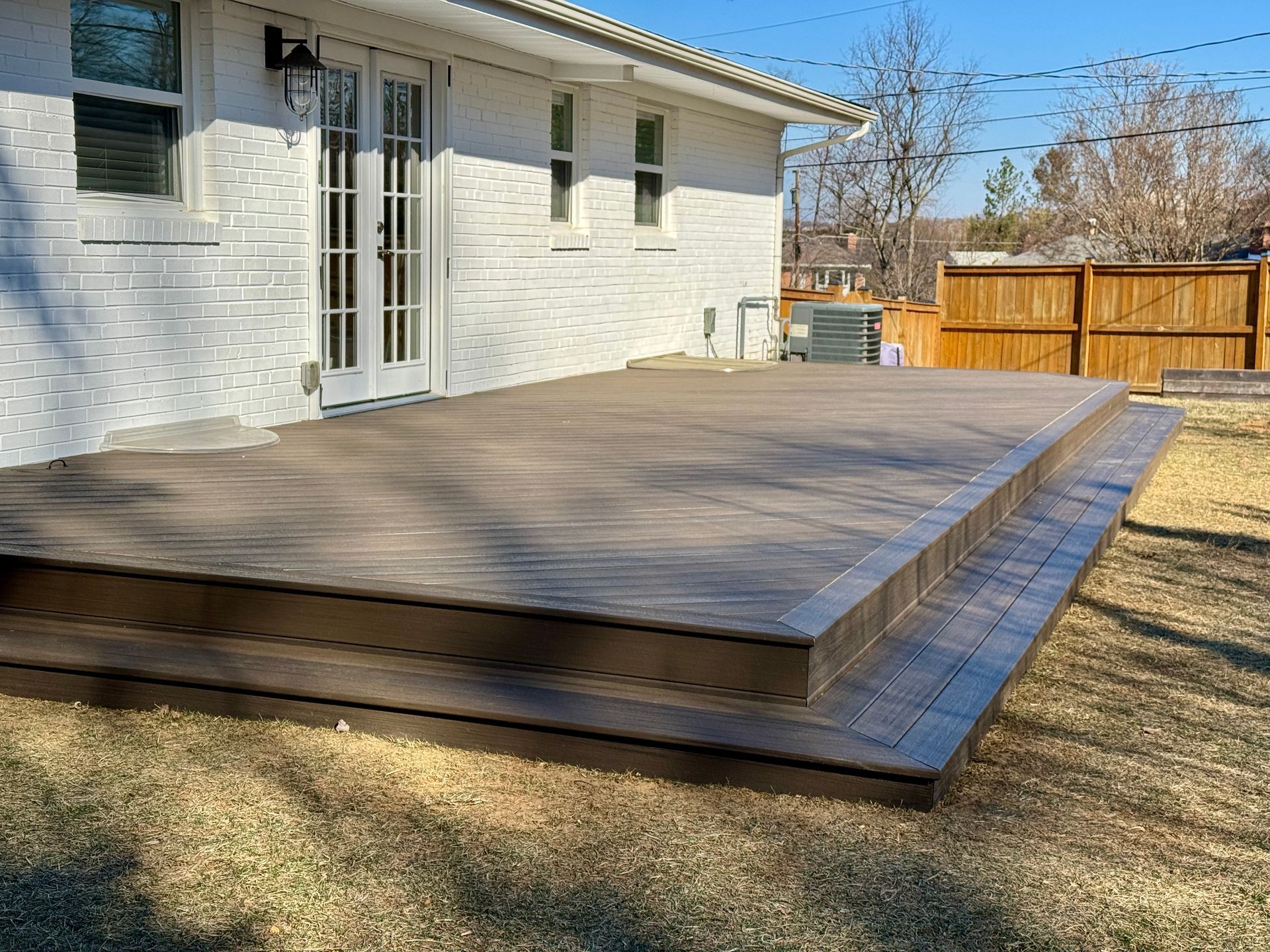 Wooden deck with steps adjacent to a white brick house, viewed from the backyard on a sunny day.