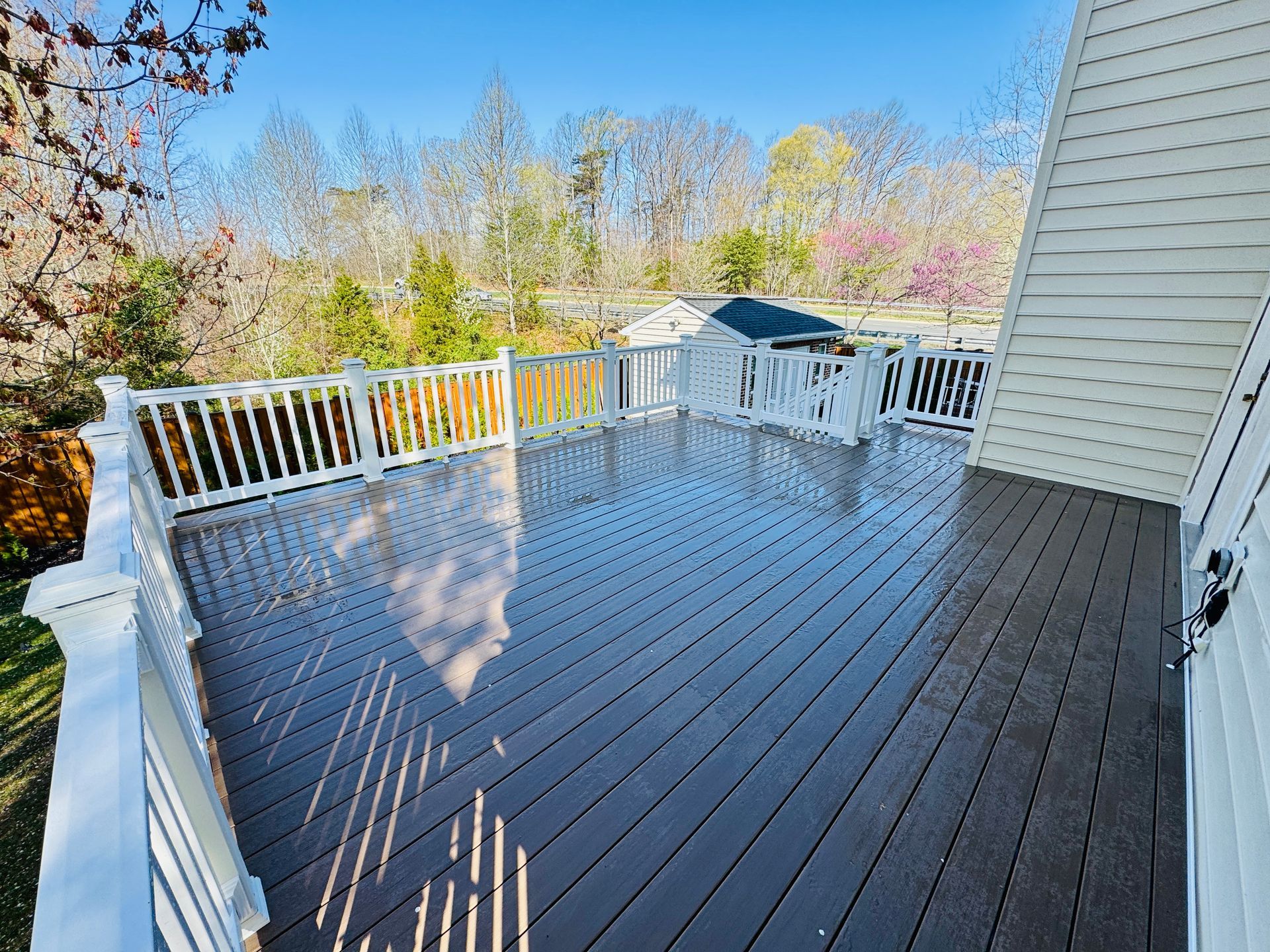 Large wooden deck with white railing, overlooking a yard and trees under a clear blue sky.