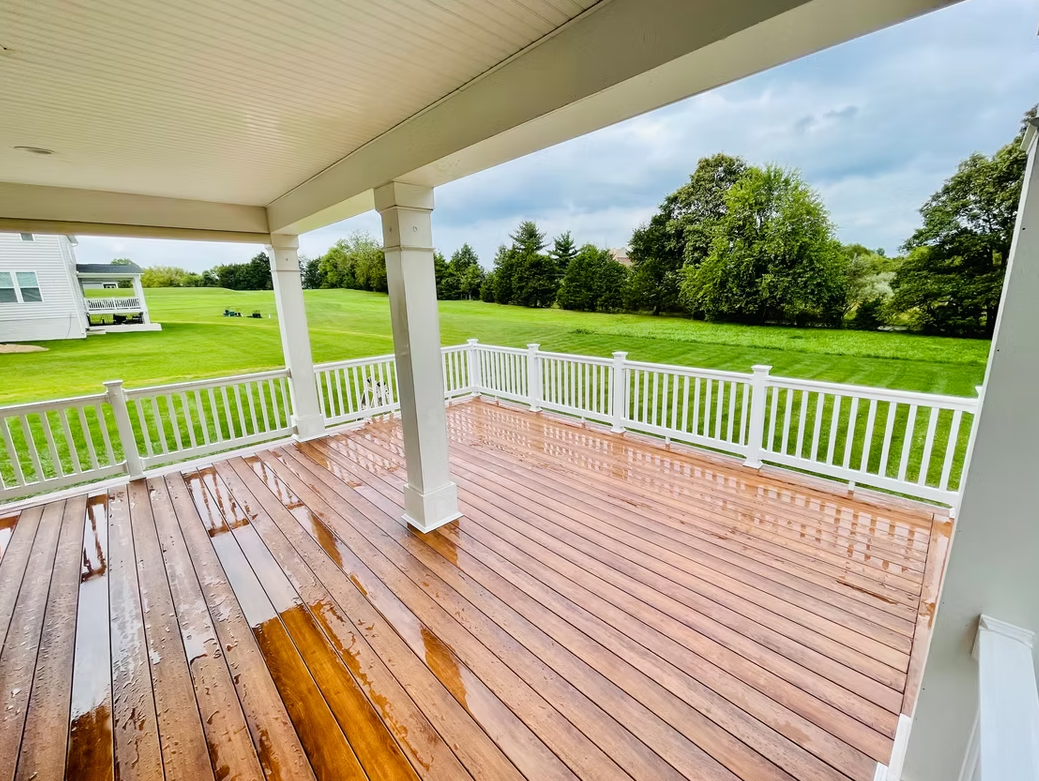 Wooden deck with white railings and columns, overlooking a green yard and trees under a cloudy sky.