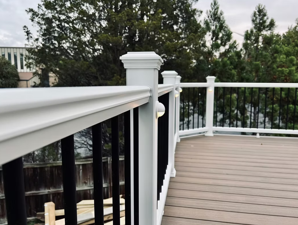 White deck railing with black balusters; brown deck; trees in background.