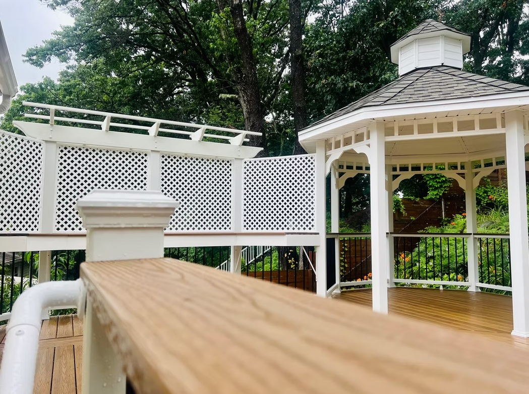Wooden deck with a white gazebo and lattice against a backdrop of green trees.