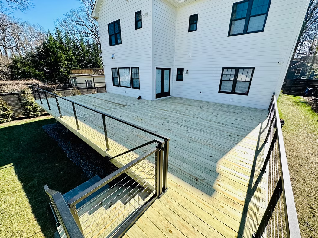 Two-story white house with large wooden deck. Black railings with glass panels. Sunny outdoor setting.