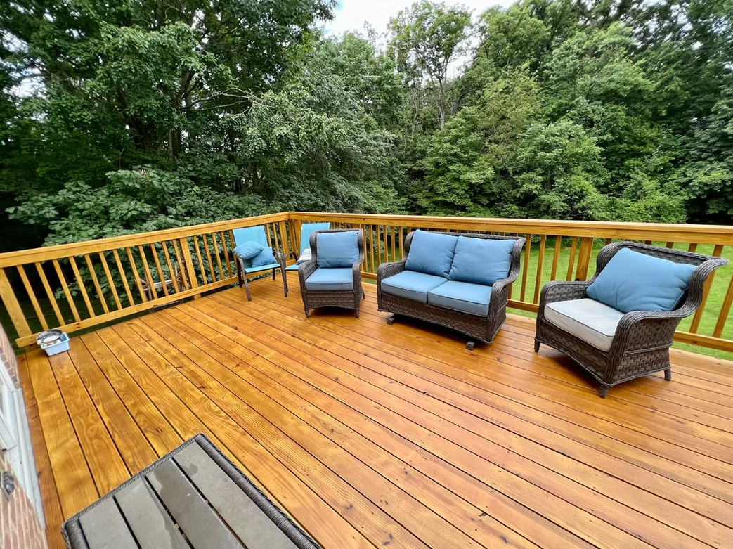 Wooden deck with outdoor furniture, overlooking a wooded area. Blue cushions on wicker seating.