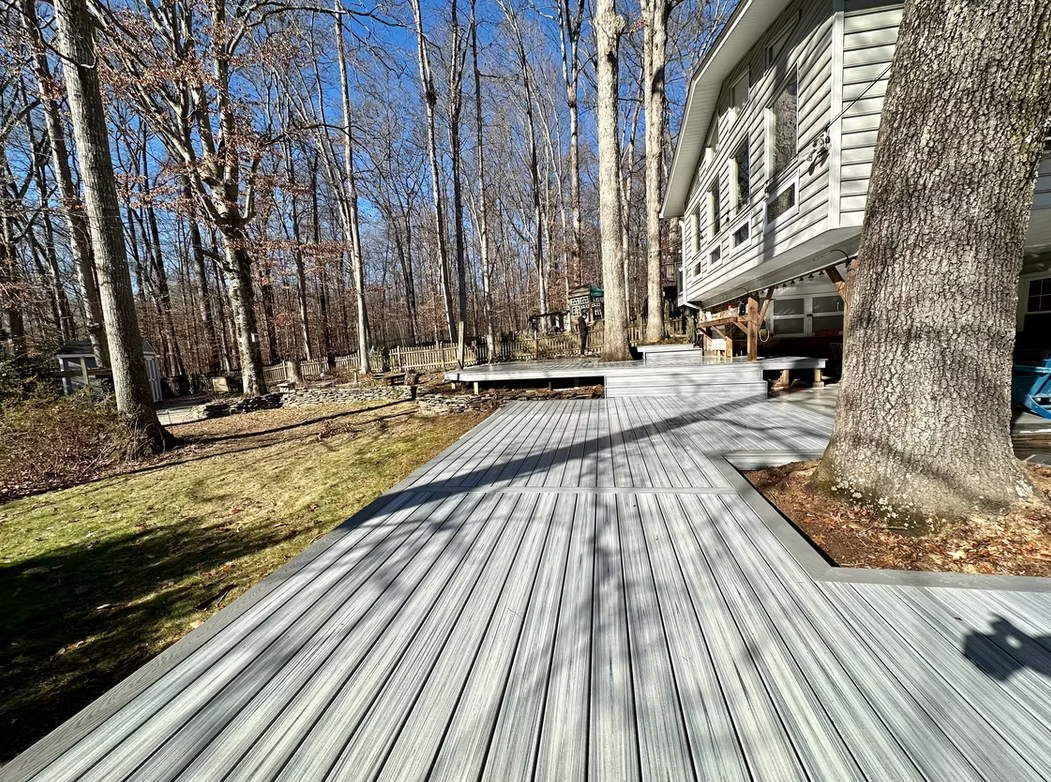 Gray composite deck extends from a white house into a wooded area on a sunny day.