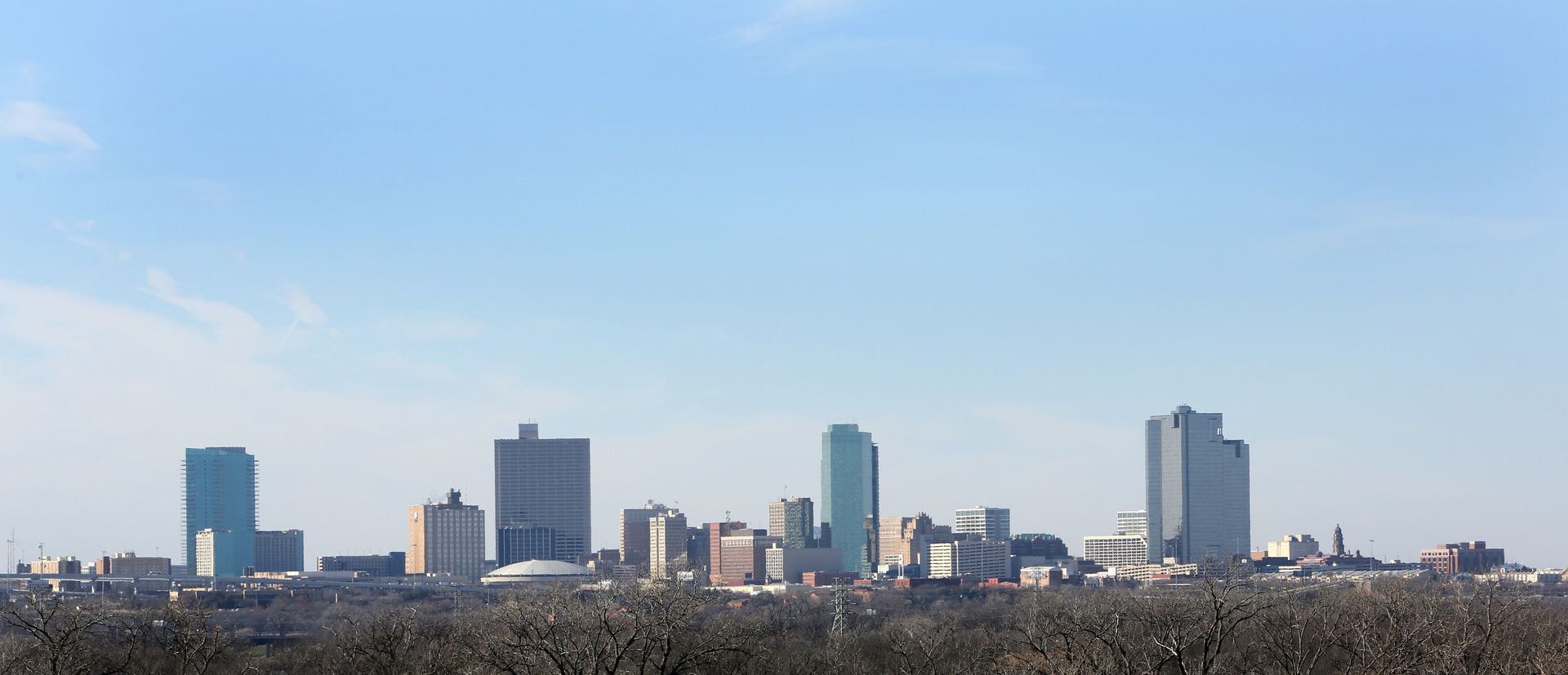 A city skyline with trees in the foreground and a blue sky in the background.