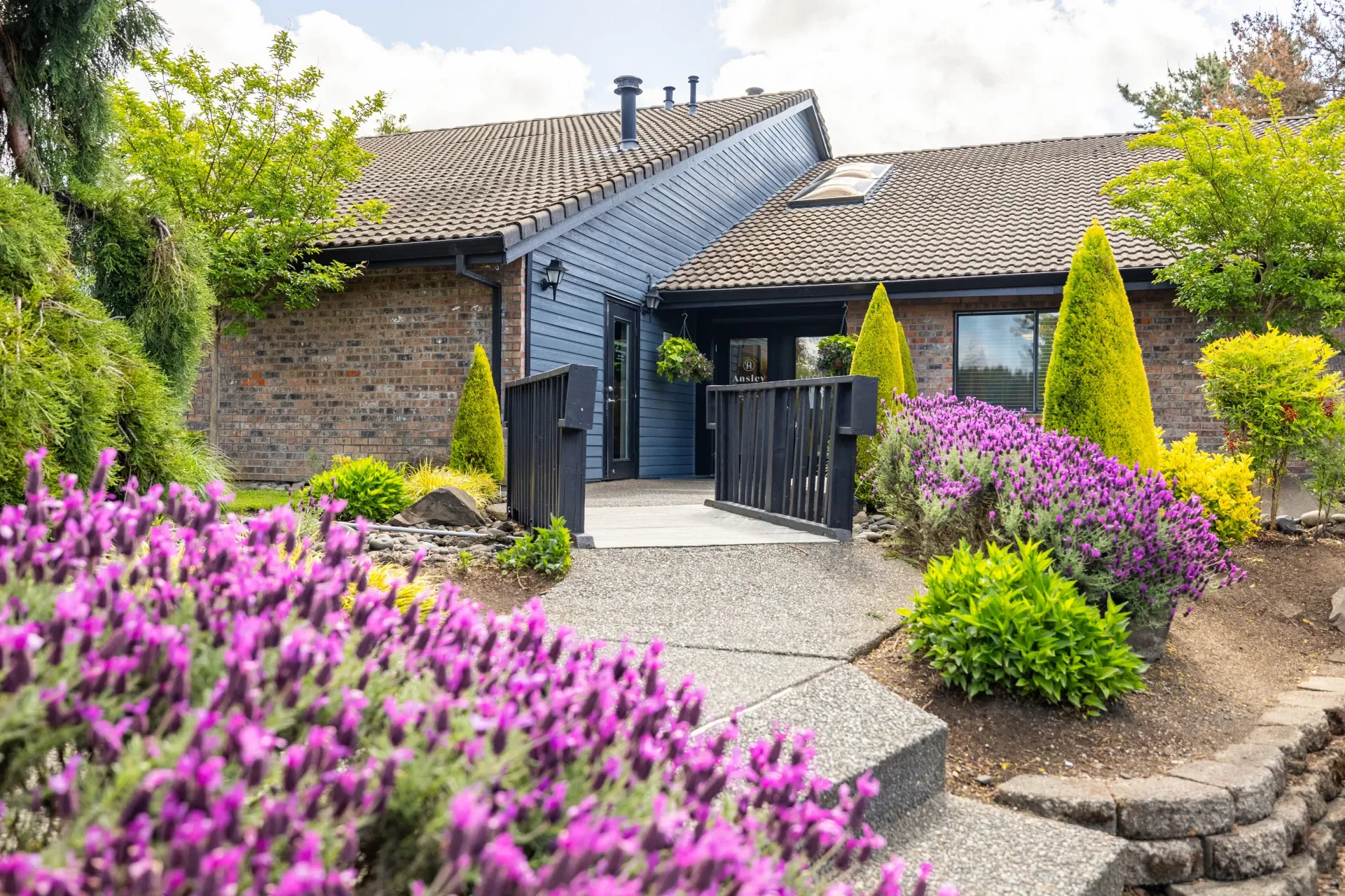 Exterior entrance to a brick multifamily building with a ramp and vibrant landscaping at Ansley Murrayhill in Tigard, OR.
