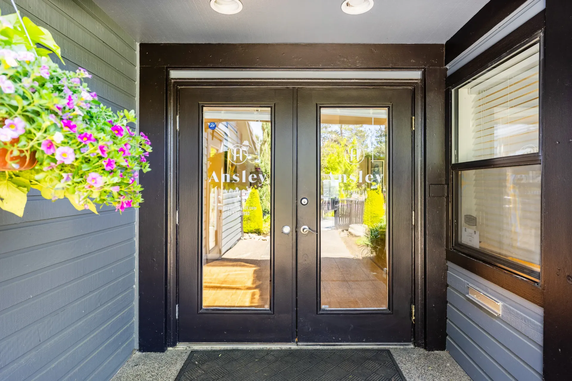 Double black glass doors with 'Ansley' signage at an apartment building entrance; flowers on the left at Ansley Murrayhill in Tigard, OR.