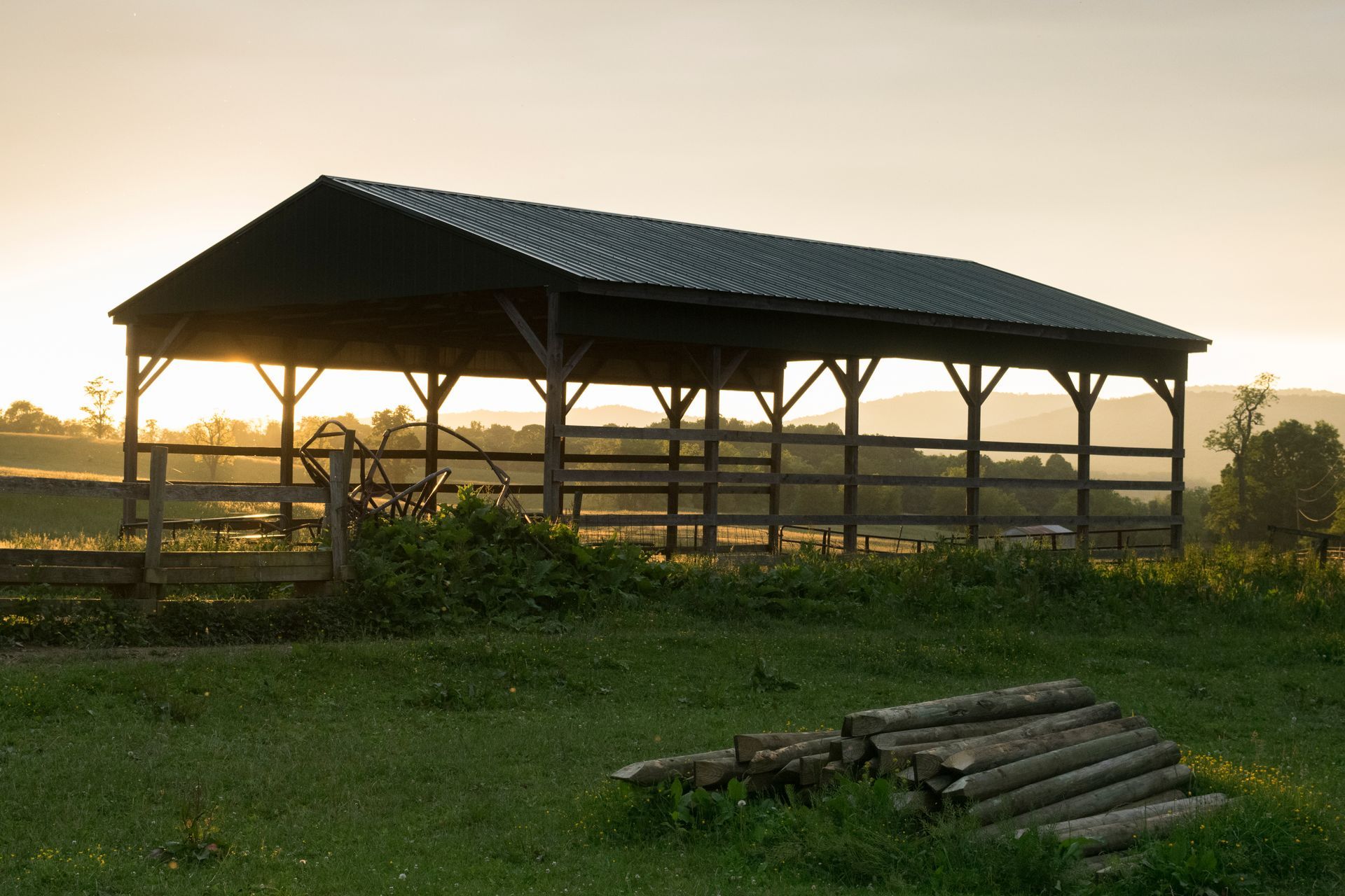 Wooden shelter in a grassy field with a fence. Logs in the foreground, mountains and sunset in the background.
