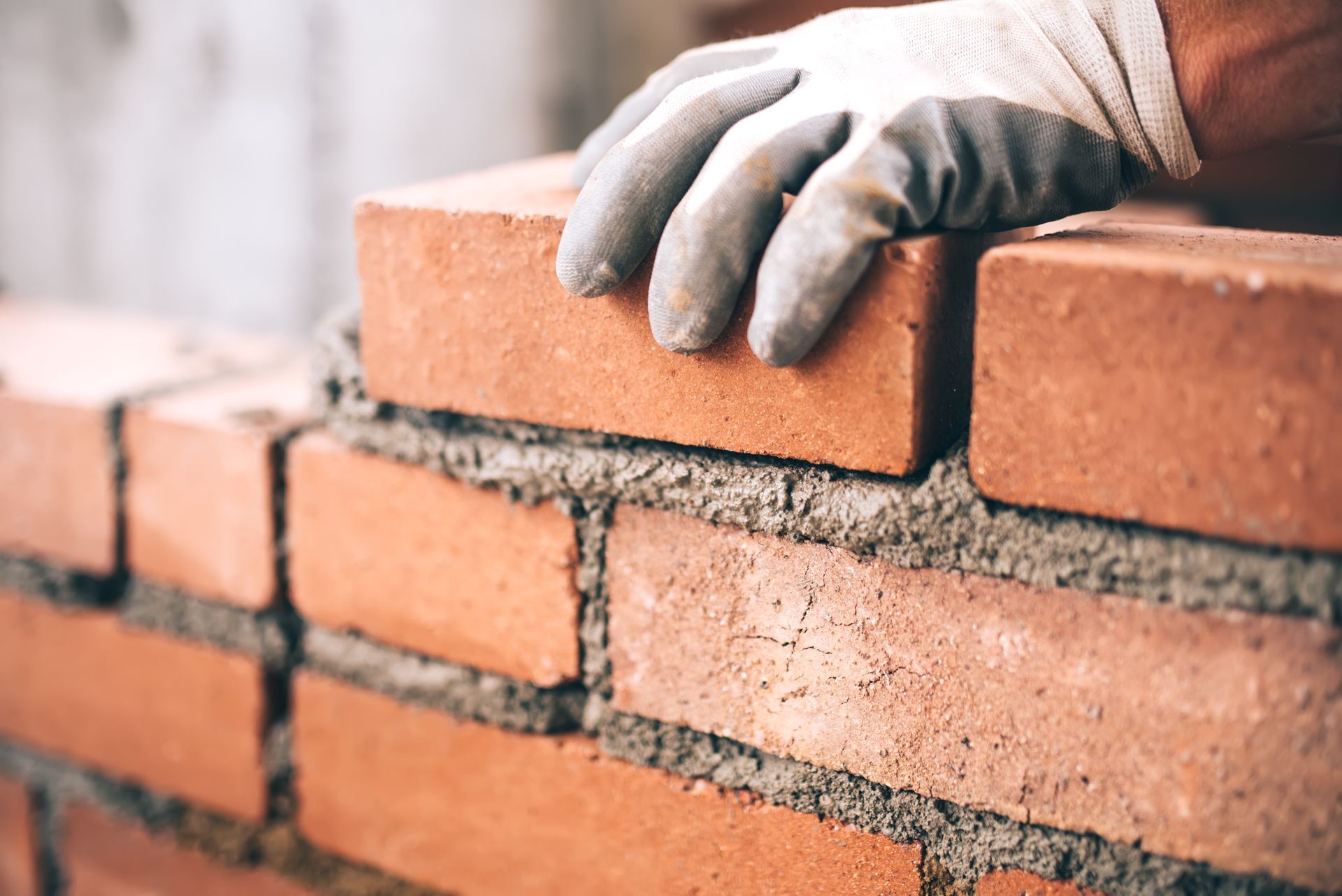 Bricklayer's gloved hand placing a red brick on a mortar-covered brick wall.