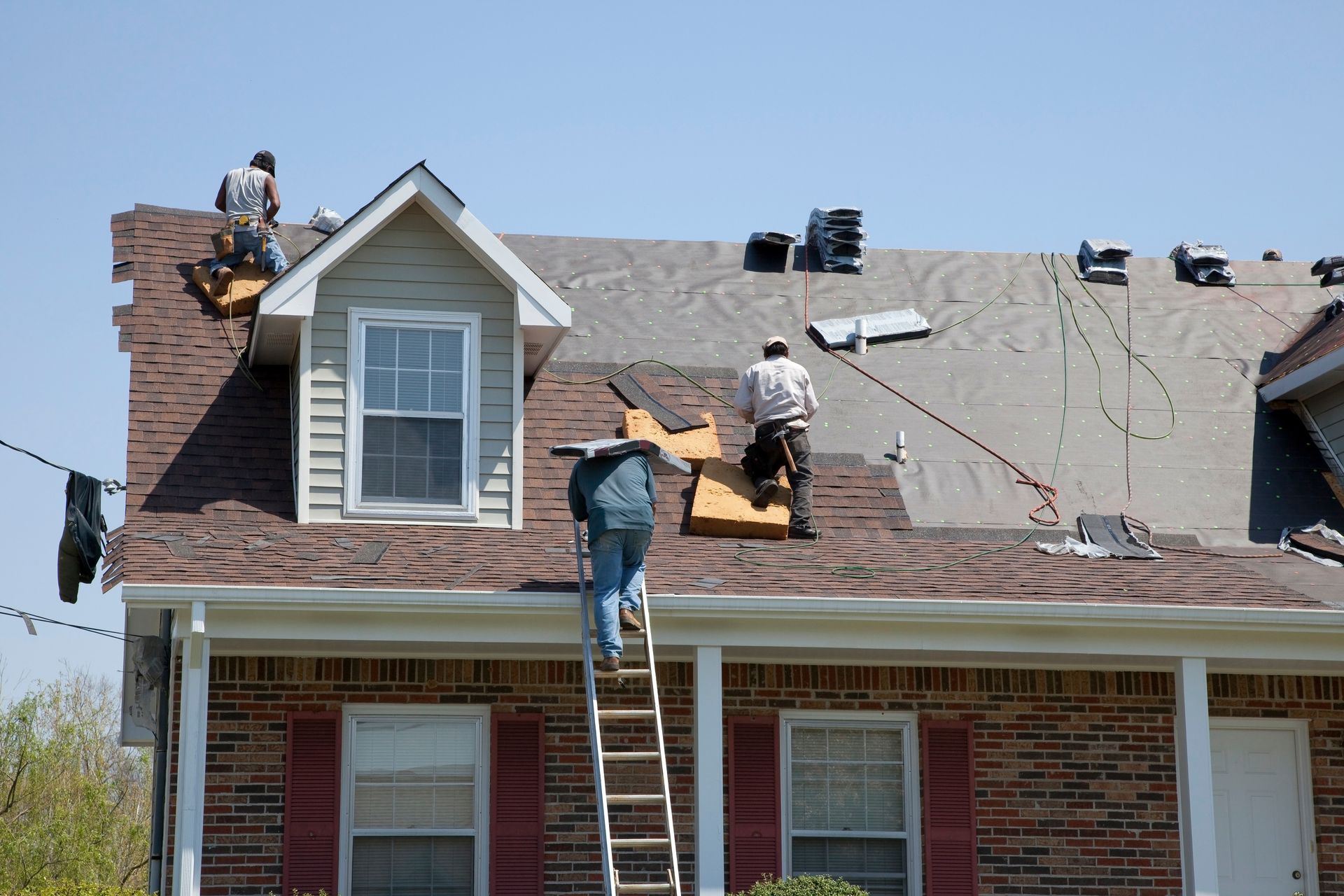 Roofers on a house roof replacing shingles. One climbs a ladder, others work with tools.