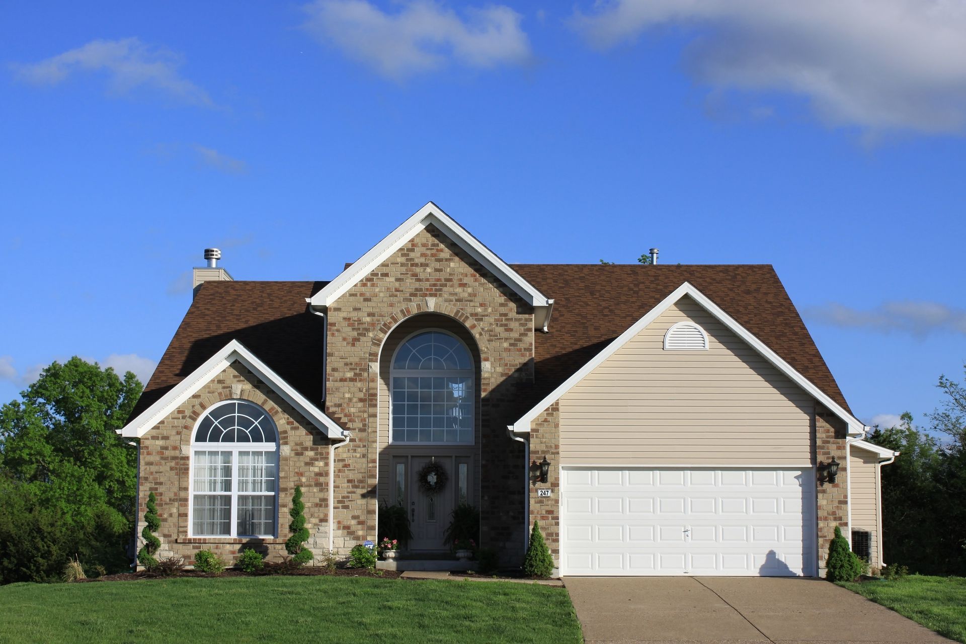 Two-story house with stone facade, beige siding, brown roof, and white garage door. Green lawn, blue sky.