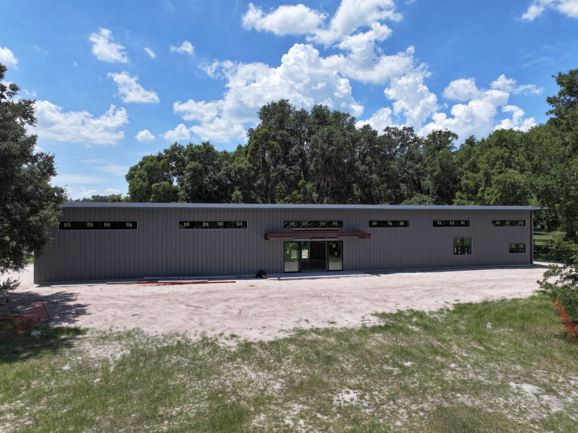 Gray industrial building with small windows under a cloudy blue sky. Surrounded by trees and a dirt lot.