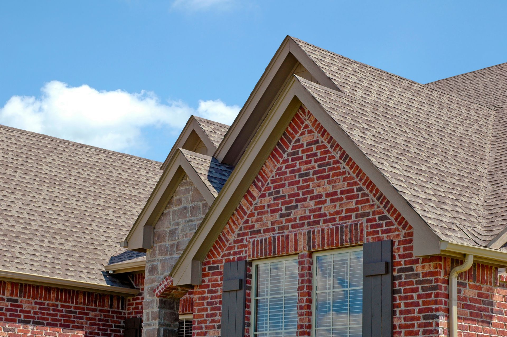 Brick house with brown shingle roof, tan trim, and blue sky.