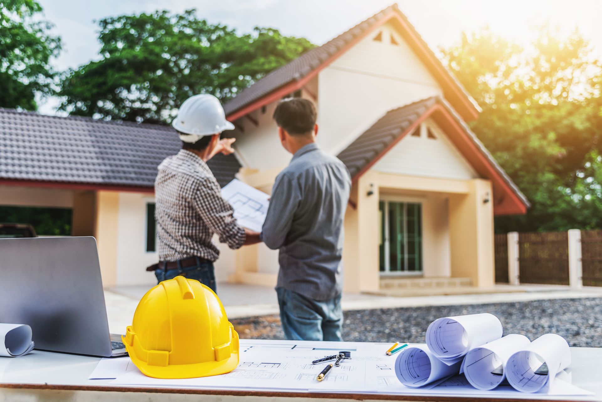 Two people in front of a house looking at blueprints; hard hat, plans and laptop on table.