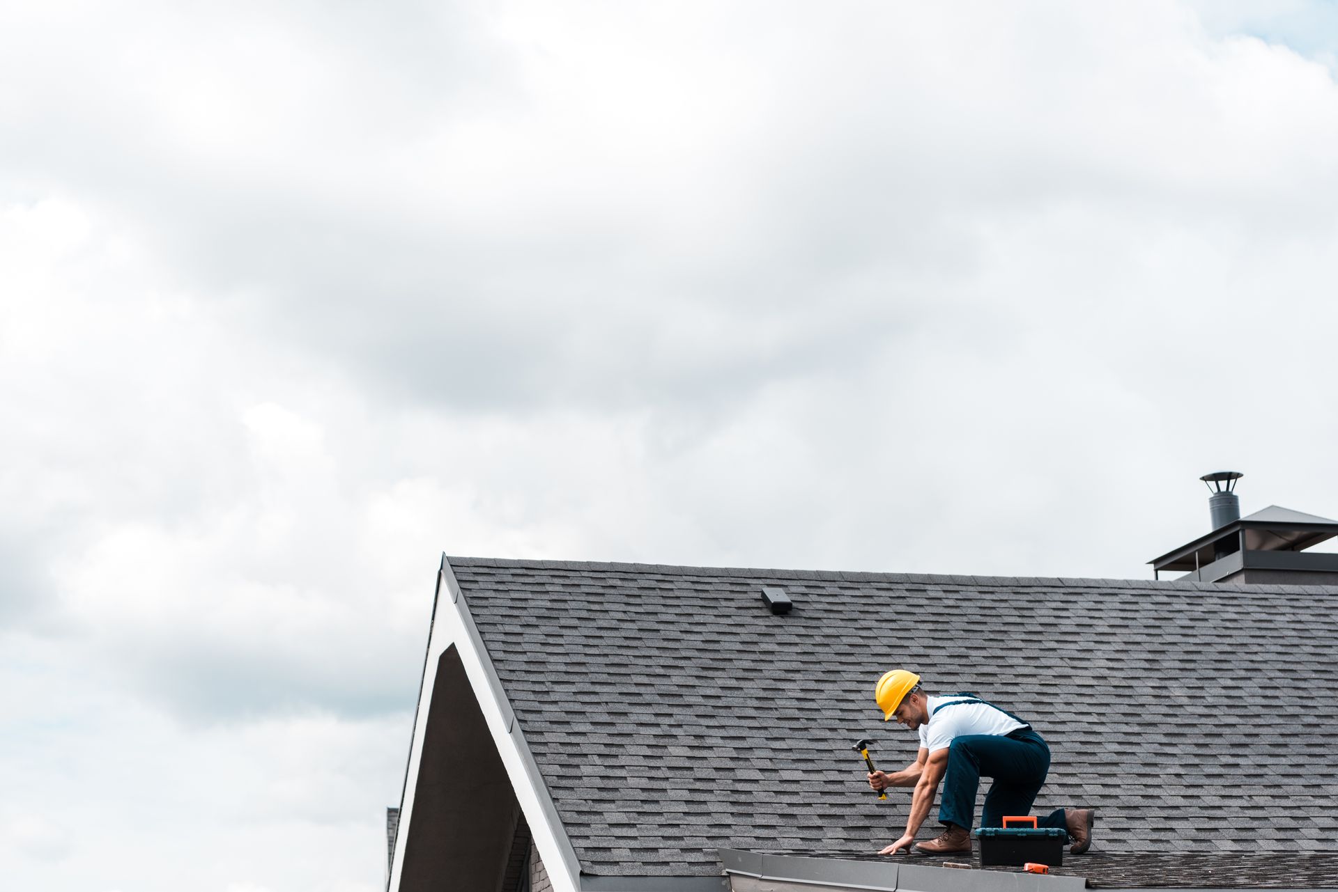 Roofer in a yellow helmet hammering shingles on a dark gray roof under a cloudy sky.