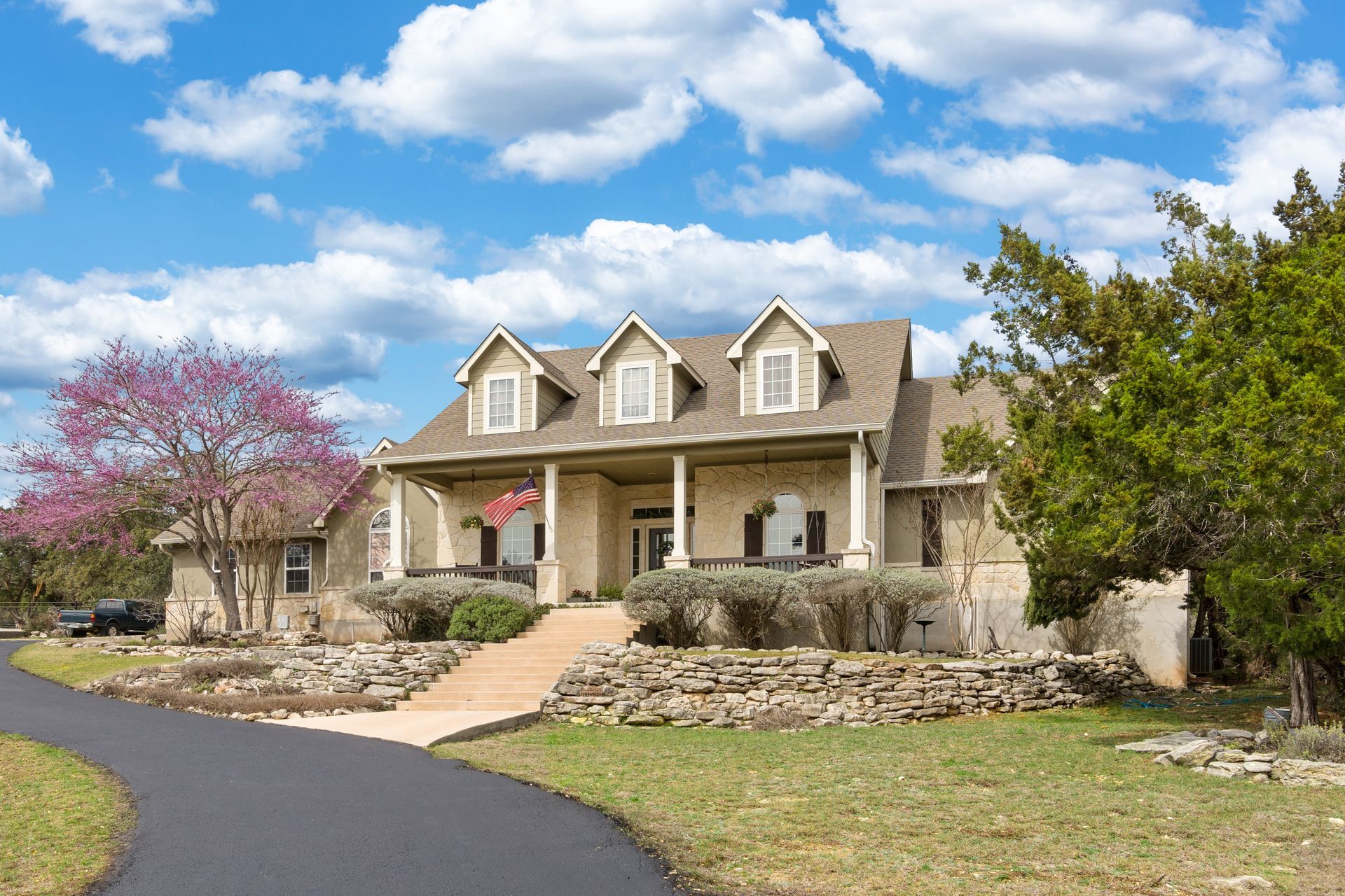 Stone house with covered porch, dormers, and curved driveway under blue sky.