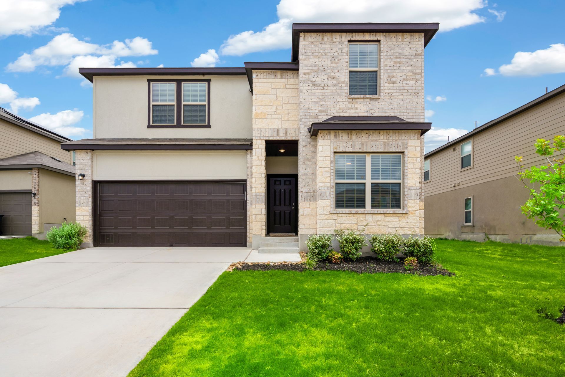 Two-story house with tan stucco and brick facade, brown garage door, and green lawn on a sunny day.