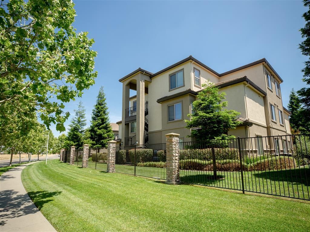 Exterior view of a modern apartment building with landscaped lawn, trees, and a black metal fence.
