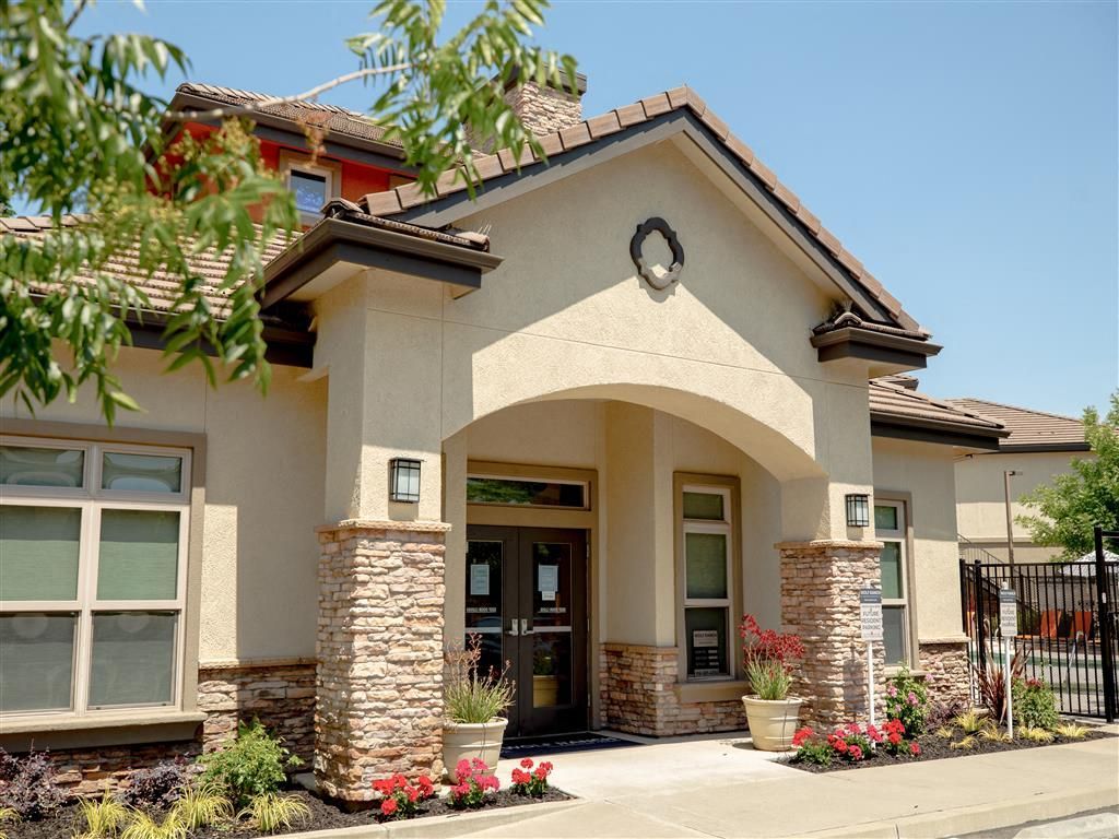 Front entrance of an apartment community featuring an arched doorway, stone pillars, and landscaped plants.