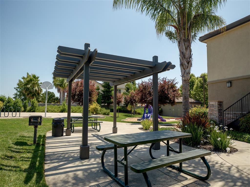 Outdoor community space with a gray pergola, picnic table, and landscaped area near a playground.