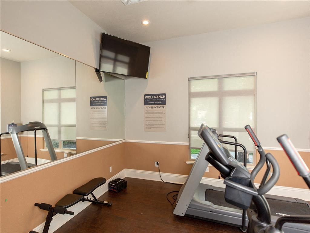 Interior of a compact community gym with treadmills, a bench, mirrors, and a wall TV.