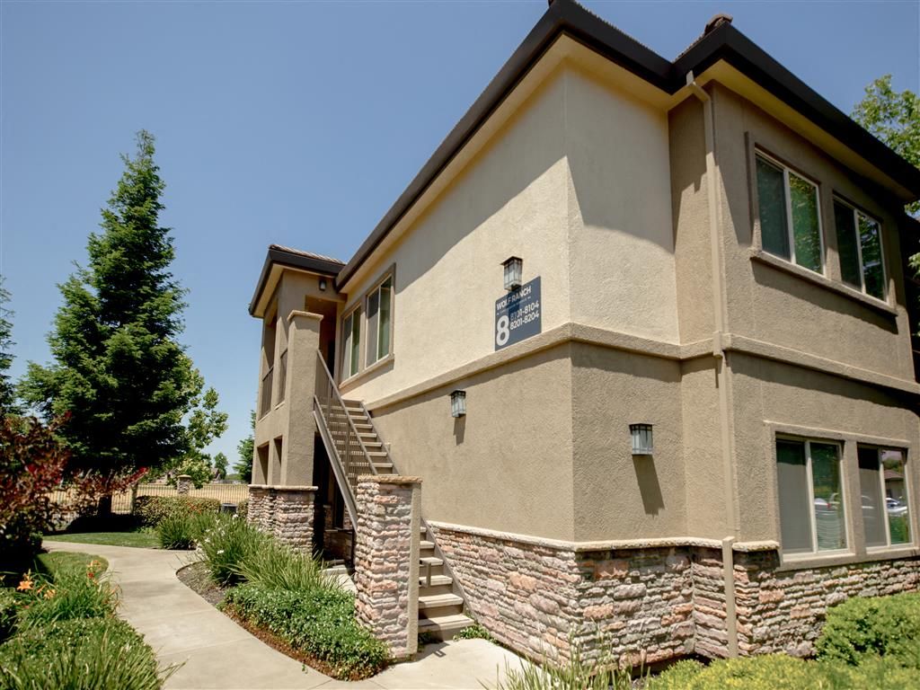 Exterior view of a two-story beige apartment building with stairs, stone accents, and surrounding landscaping.