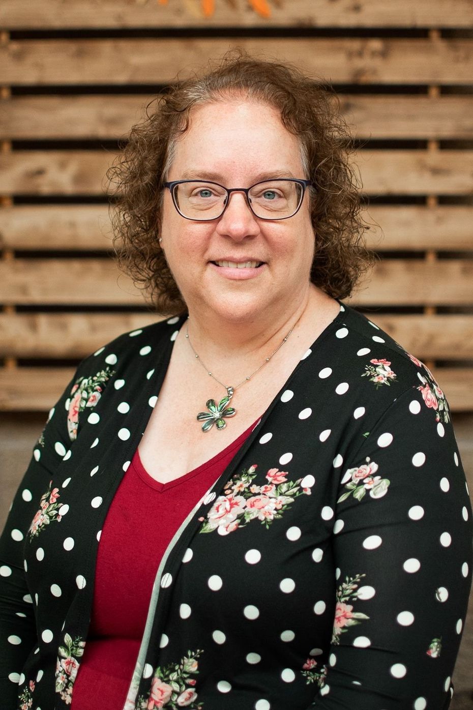A woman with curly brown hair and glasses wears a red shirt and a black floral-print cardigan, smiling against wood slats.