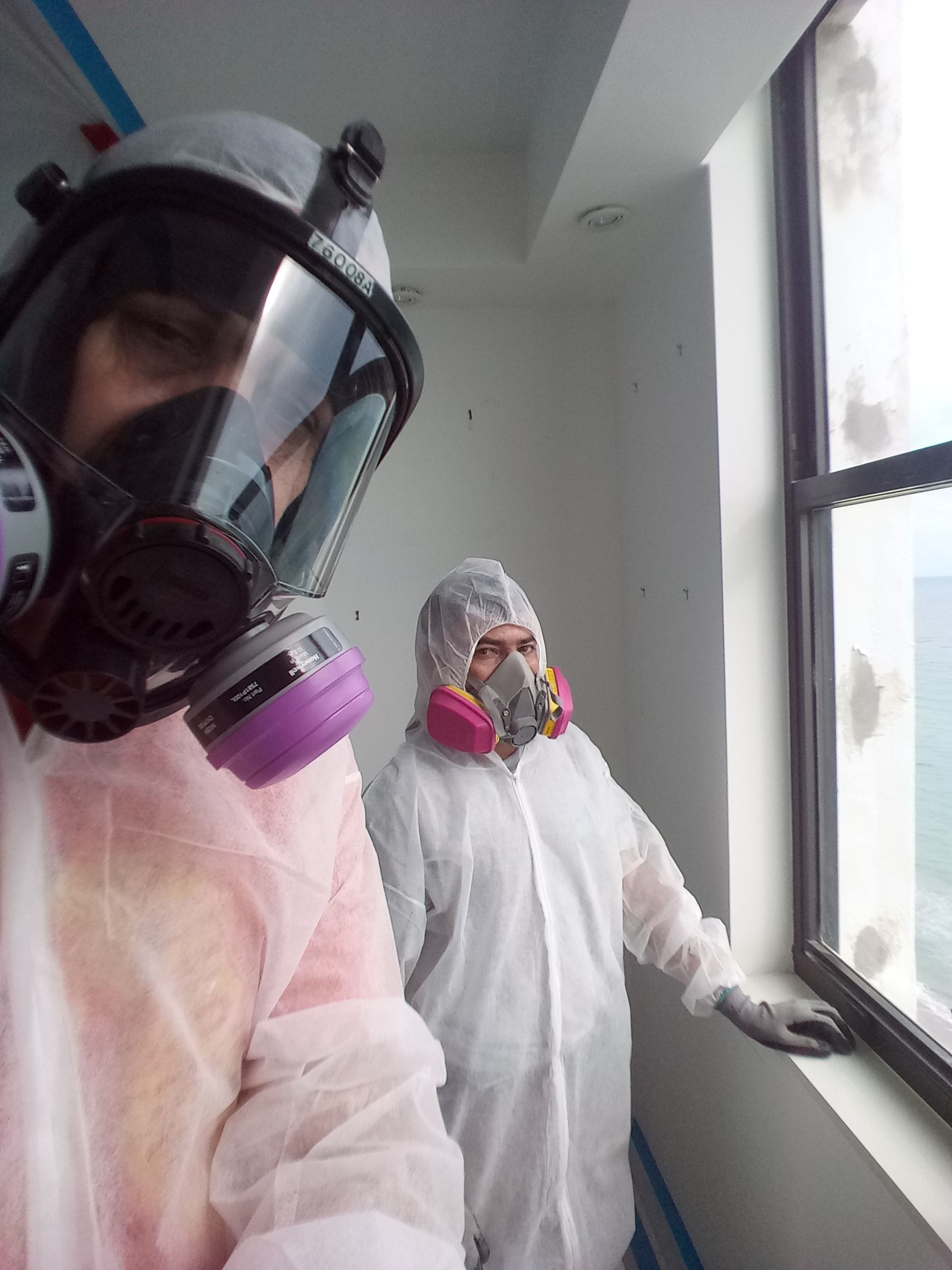 Two people wearing gas masks are looking out a window