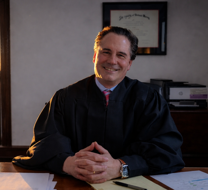 Judge smiling, wearing a black robe, seated at a desk, sunlight, framed document in background.
