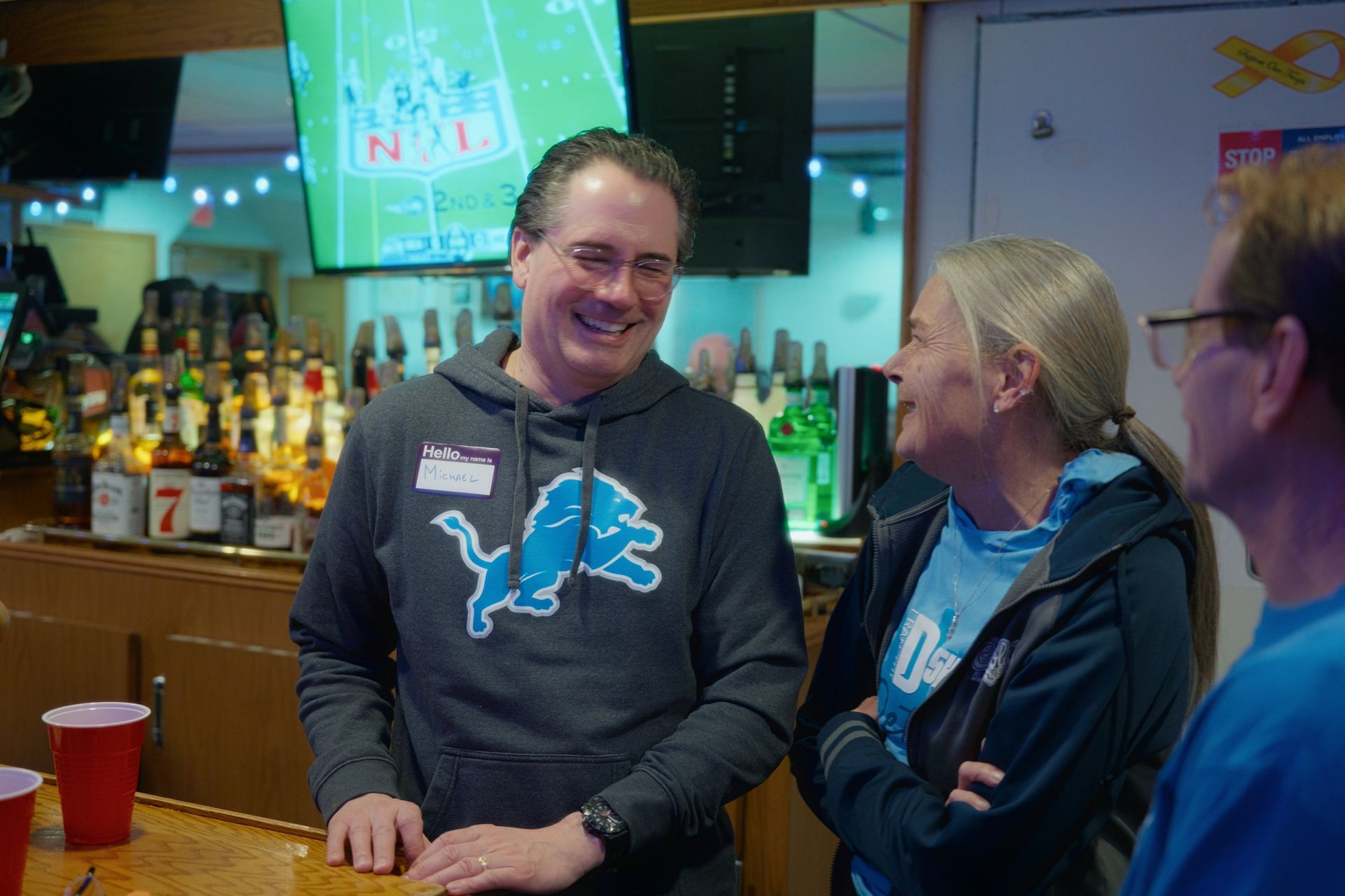 Three people smiling at a bar, Detroit Lions fans; one wears a Lions hoodie, TV screen visible in background.