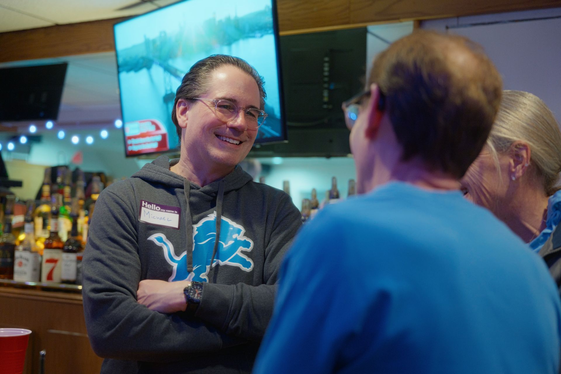 Man smiling, arms crossed, wearing a Detroit Lions sweatshirt, talking to two other people at a bar.