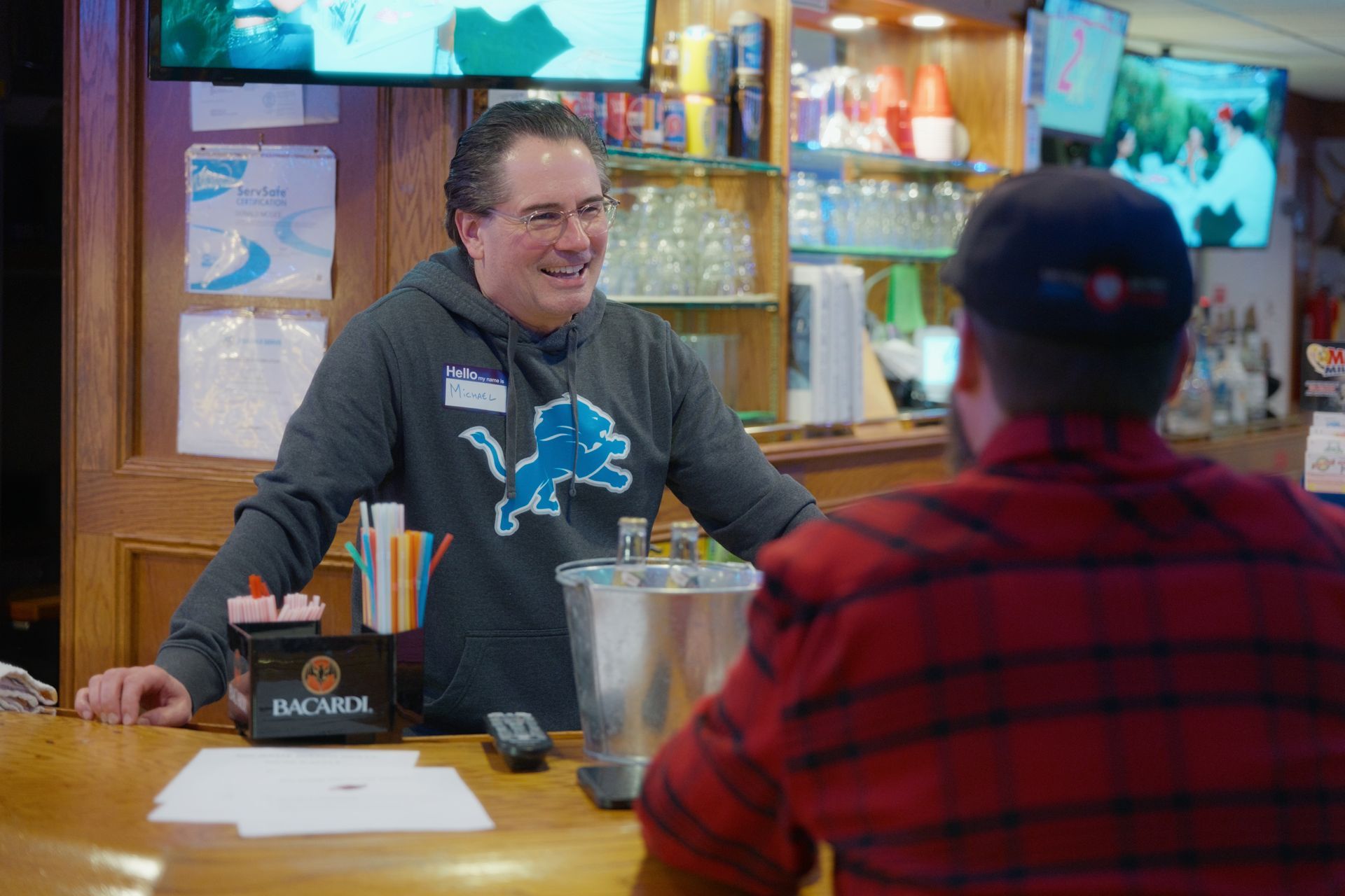 Bartender in a Detroit Lions sweatshirt smiles at a customer at a bar.