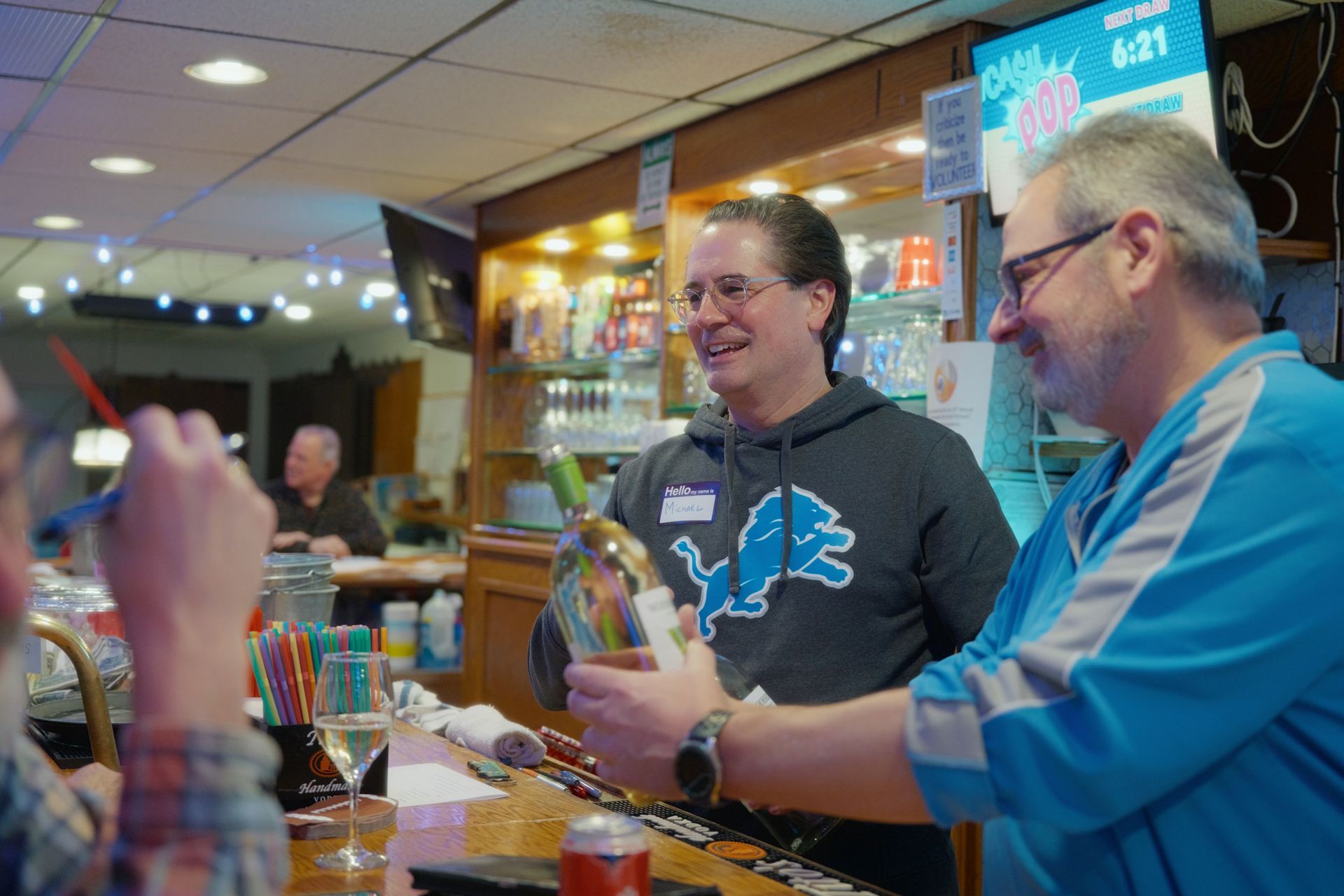 Two men at a bar, one in a Detroit Lions sweatshirt, holding a decorated bottle; bar setting.