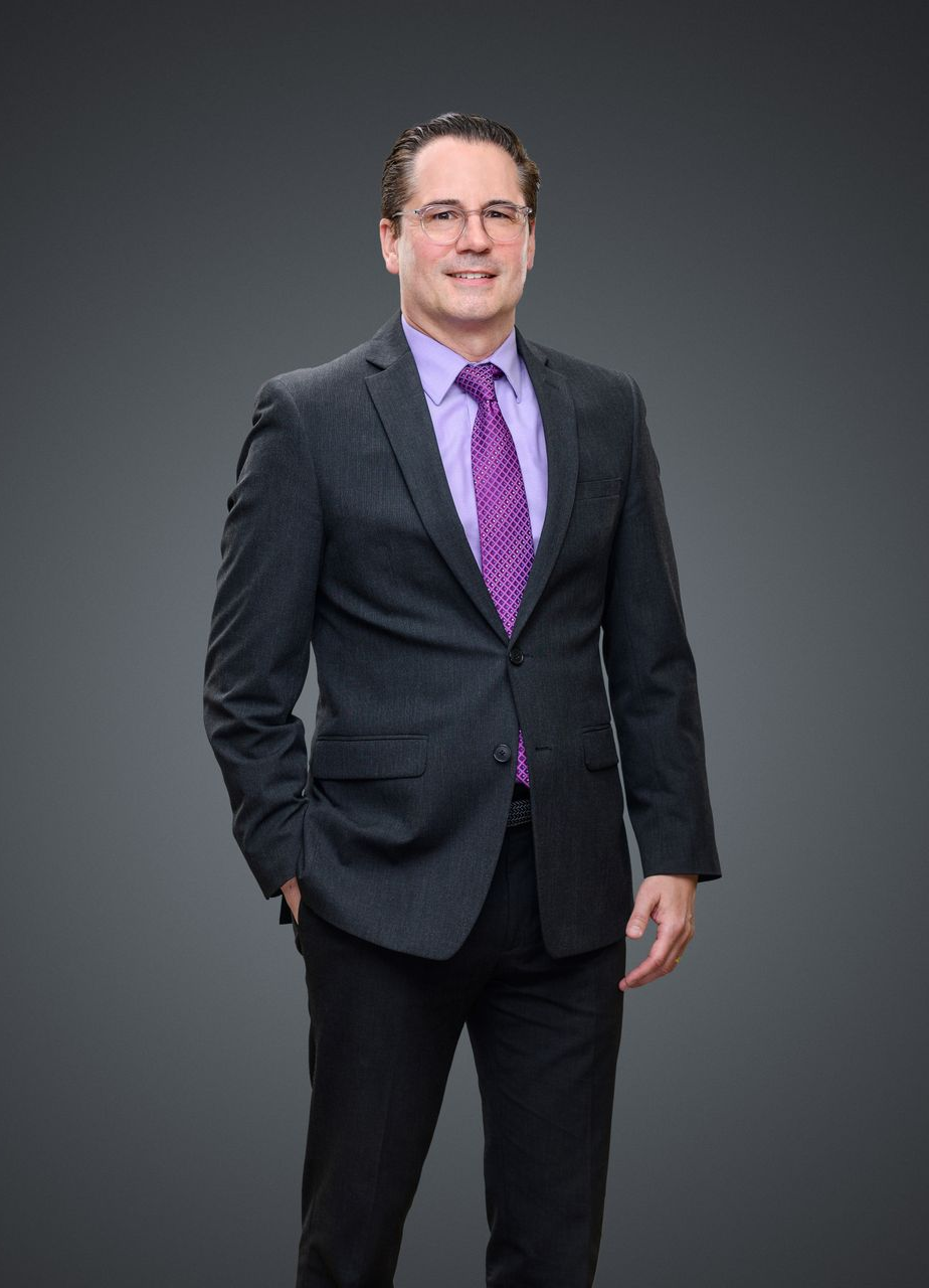 Michaelin suit, smiling, standing with hand in pocket, against a gray backdrop.