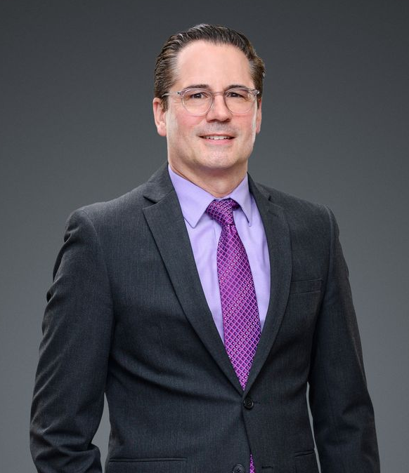 Michael in a suit smiles, wearing glasses, purple tie and shirt, against a gray backdrop.