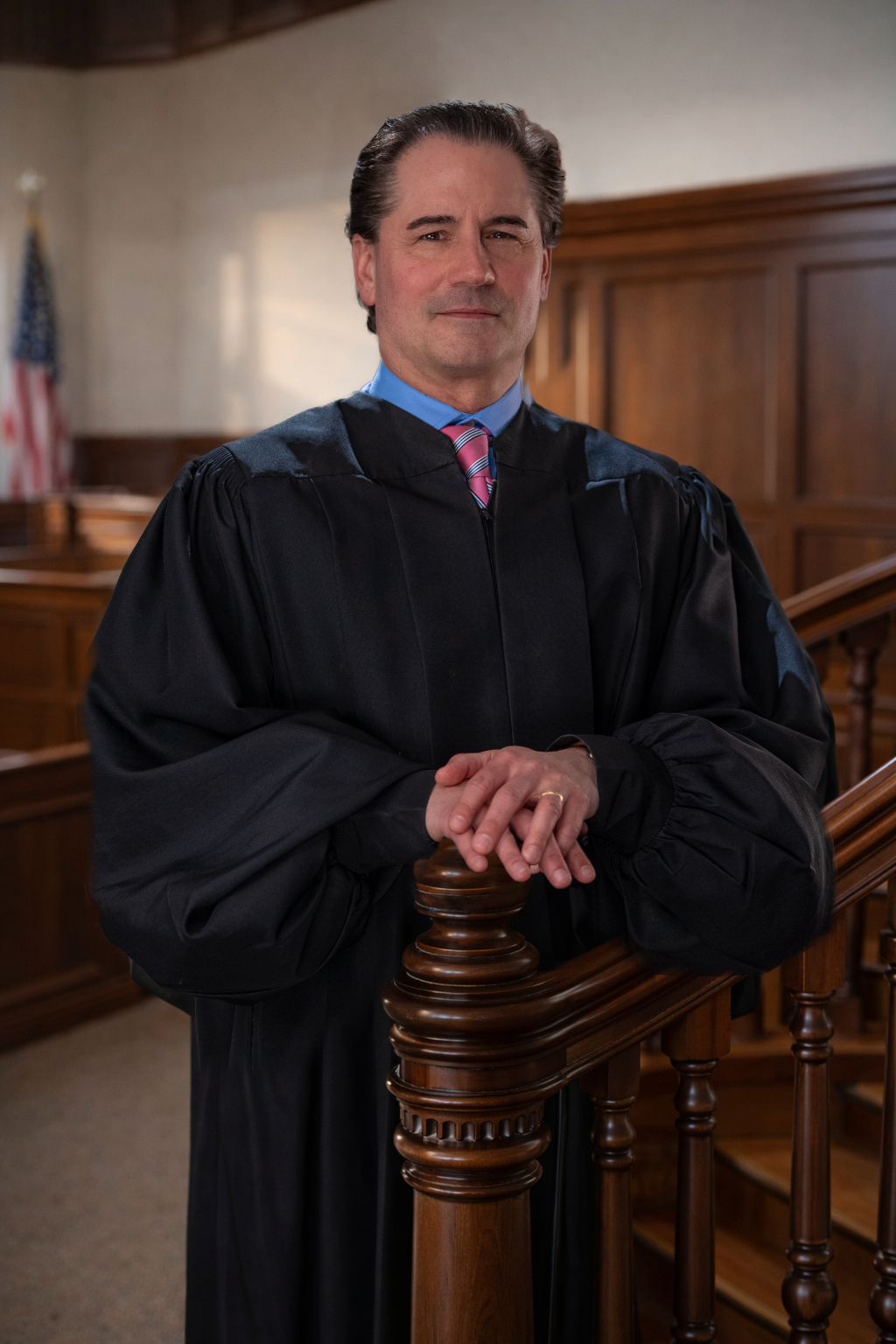Michael in black robe, leaning on wooden railing in courtroom, hands clasped.
