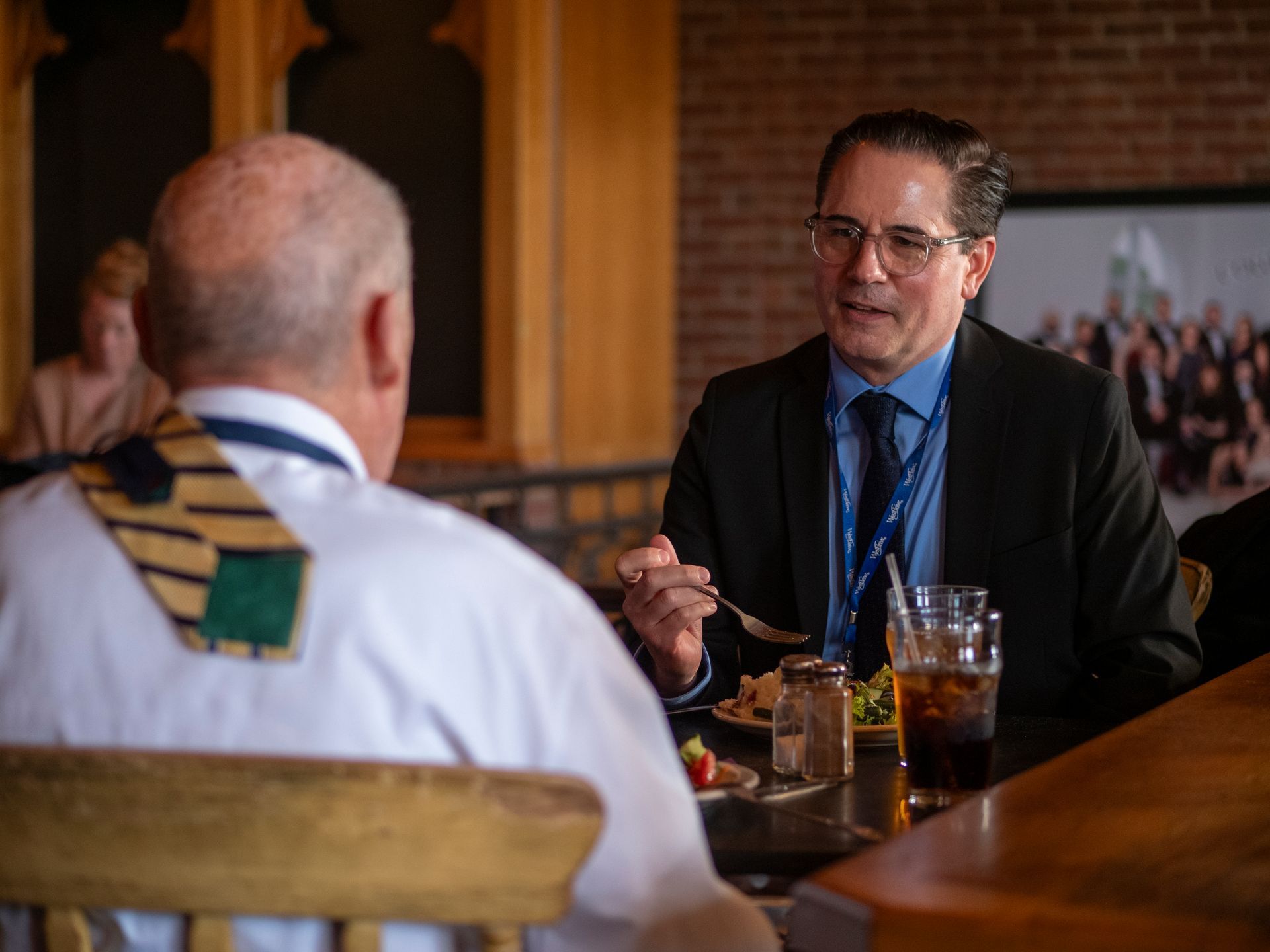 Man in suit talks to another person while eating at a table; they are indoors.
