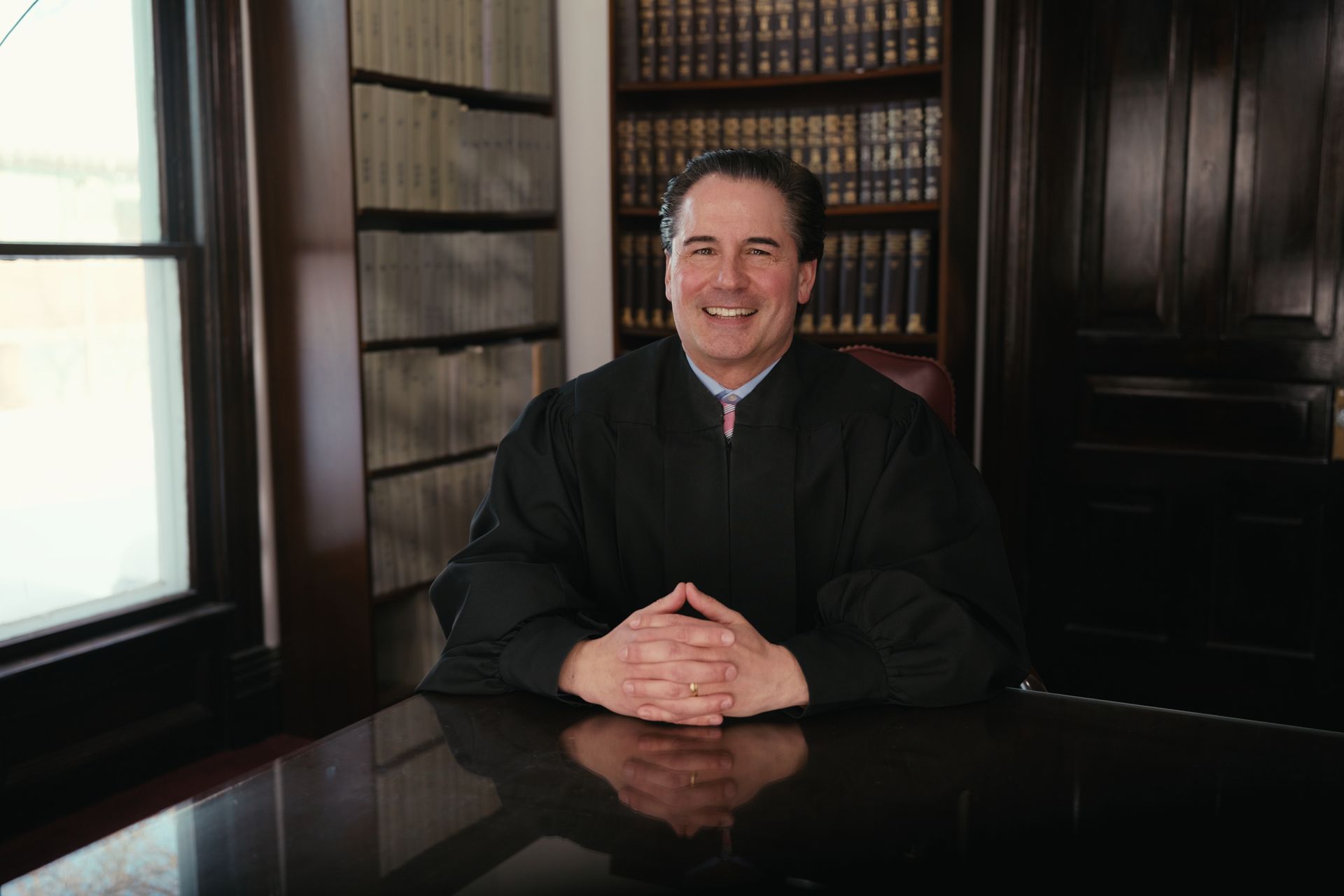 Smiling Magistrate Michael Sawicky in black robe seated at a desk, library background.