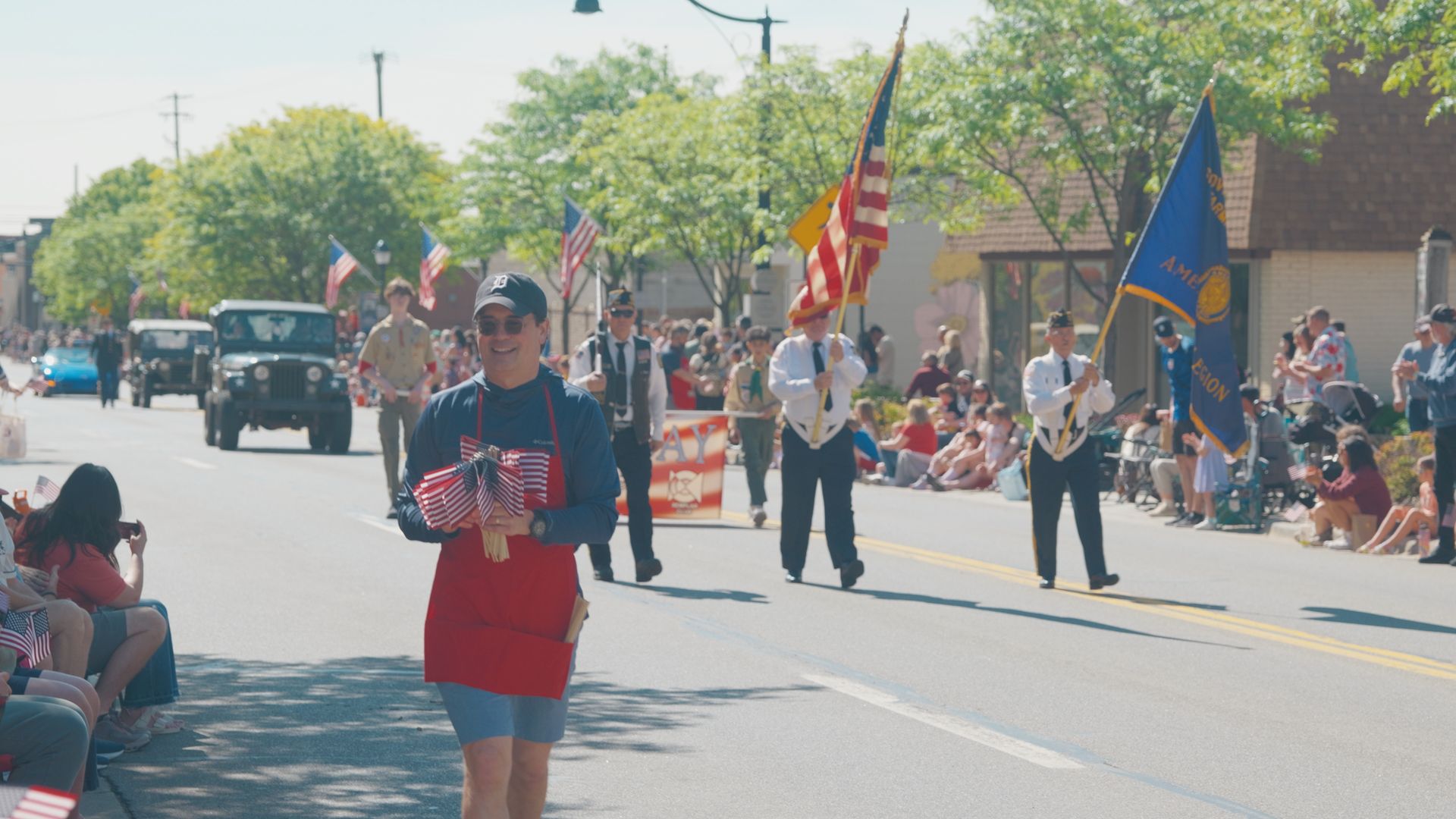 Michael Sawicky in the memorial day parade in Farmington Hills. marchers carrying flags along a sunny street, with spectators seated on both sides.
