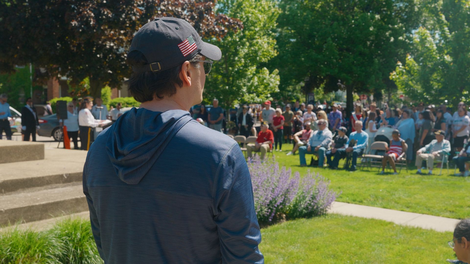 Michael Sawicky in the memorial day parade in Farmington Hills in a navy hoodie and cap stands facing a seated crowd in a sunny park near steps and flowers.