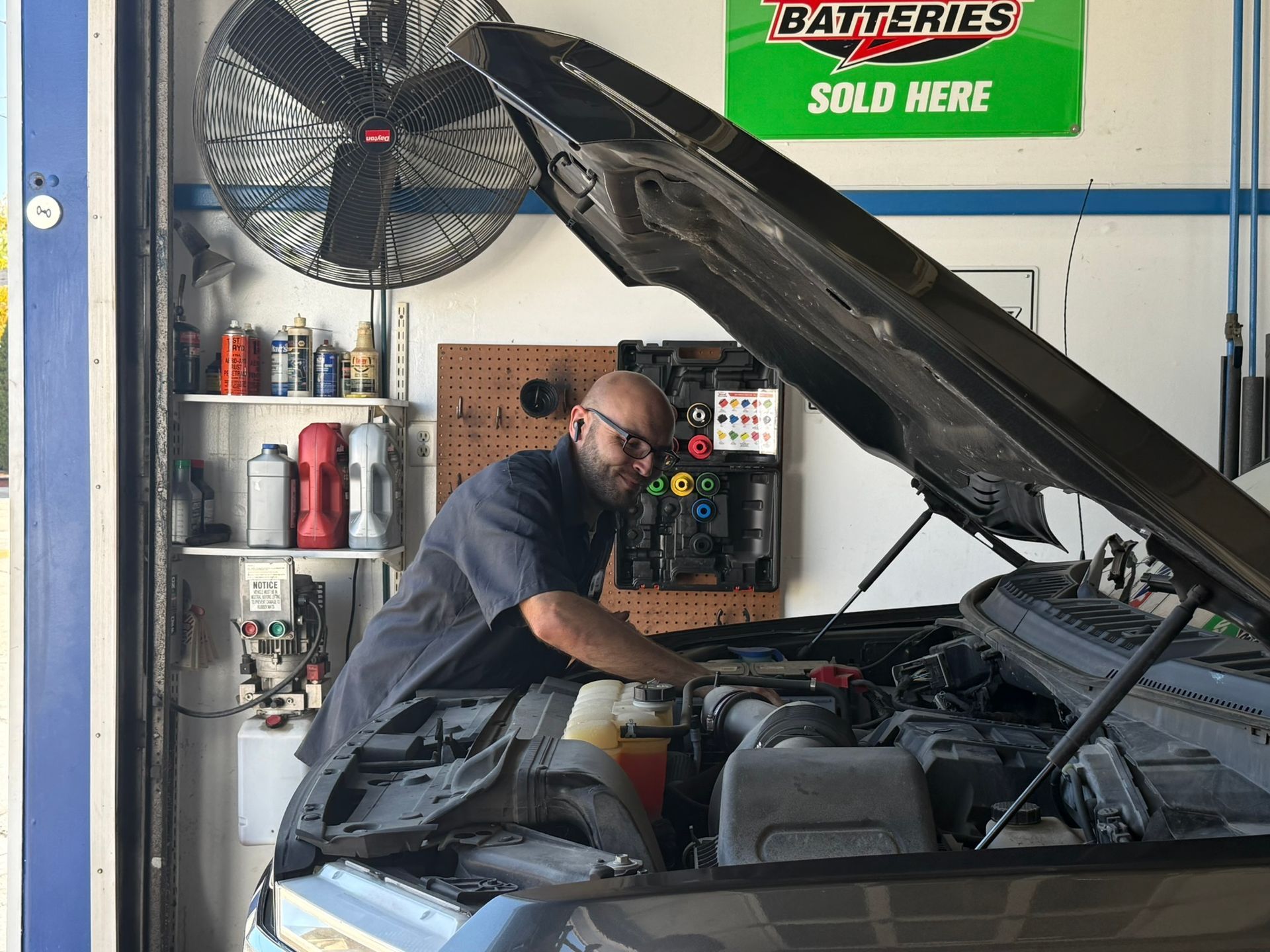 Mechanic in blue overalls inspecting engine parts in a garage with a car on a lift.