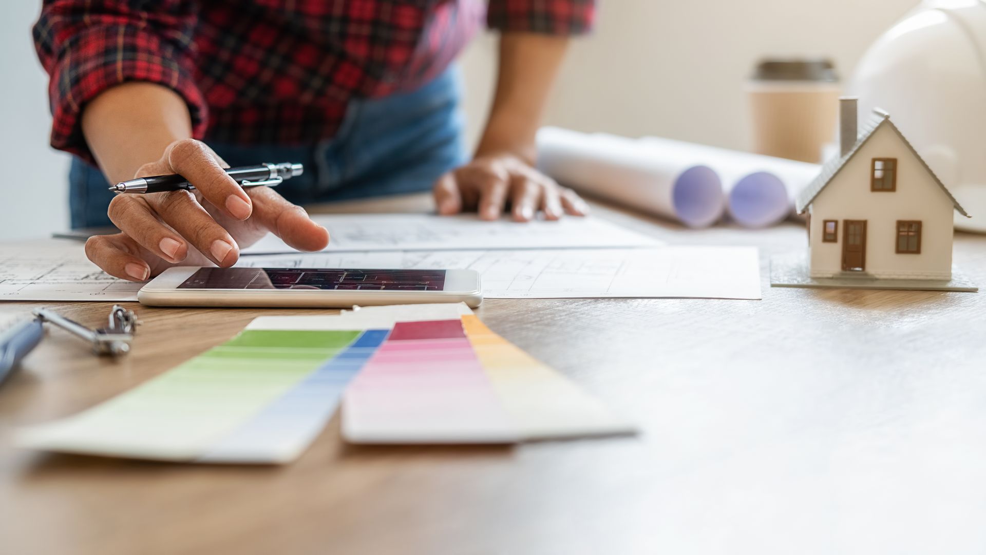 Designer using a phone over blueprints with color swatches and a small model house.