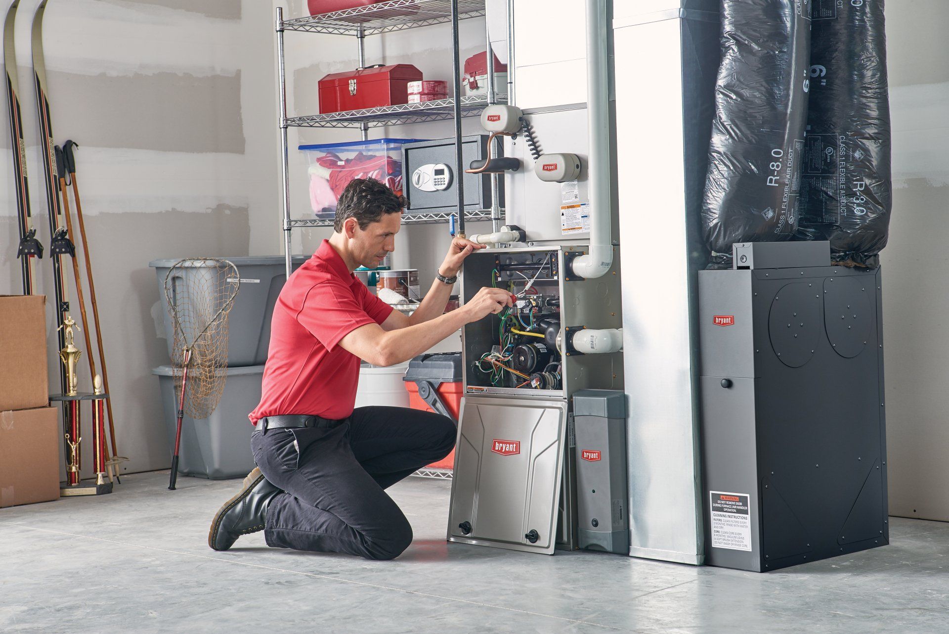 HVAC technician kneeling, inspecting furnace in a garage. Red shirt, black pants, gray and black equipment.