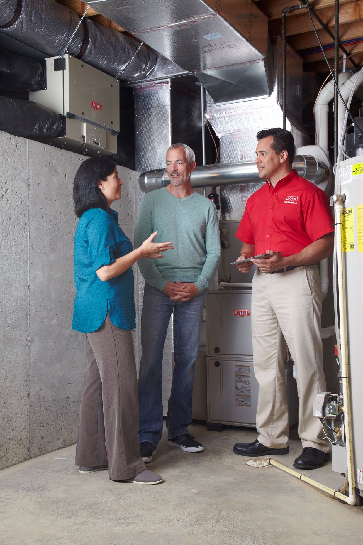 A technician in red shirt consults with a couple in a basement, discussing HVAC equipment.