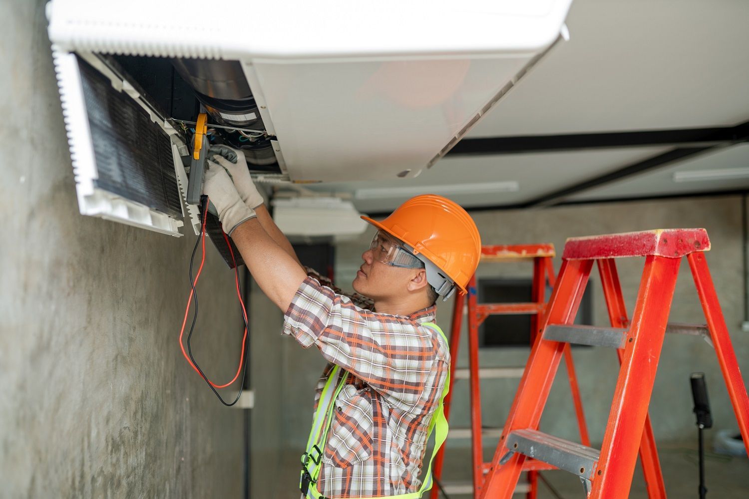 A worker repairing a ceiling air conditioning unit with tools and a red ladder nearby