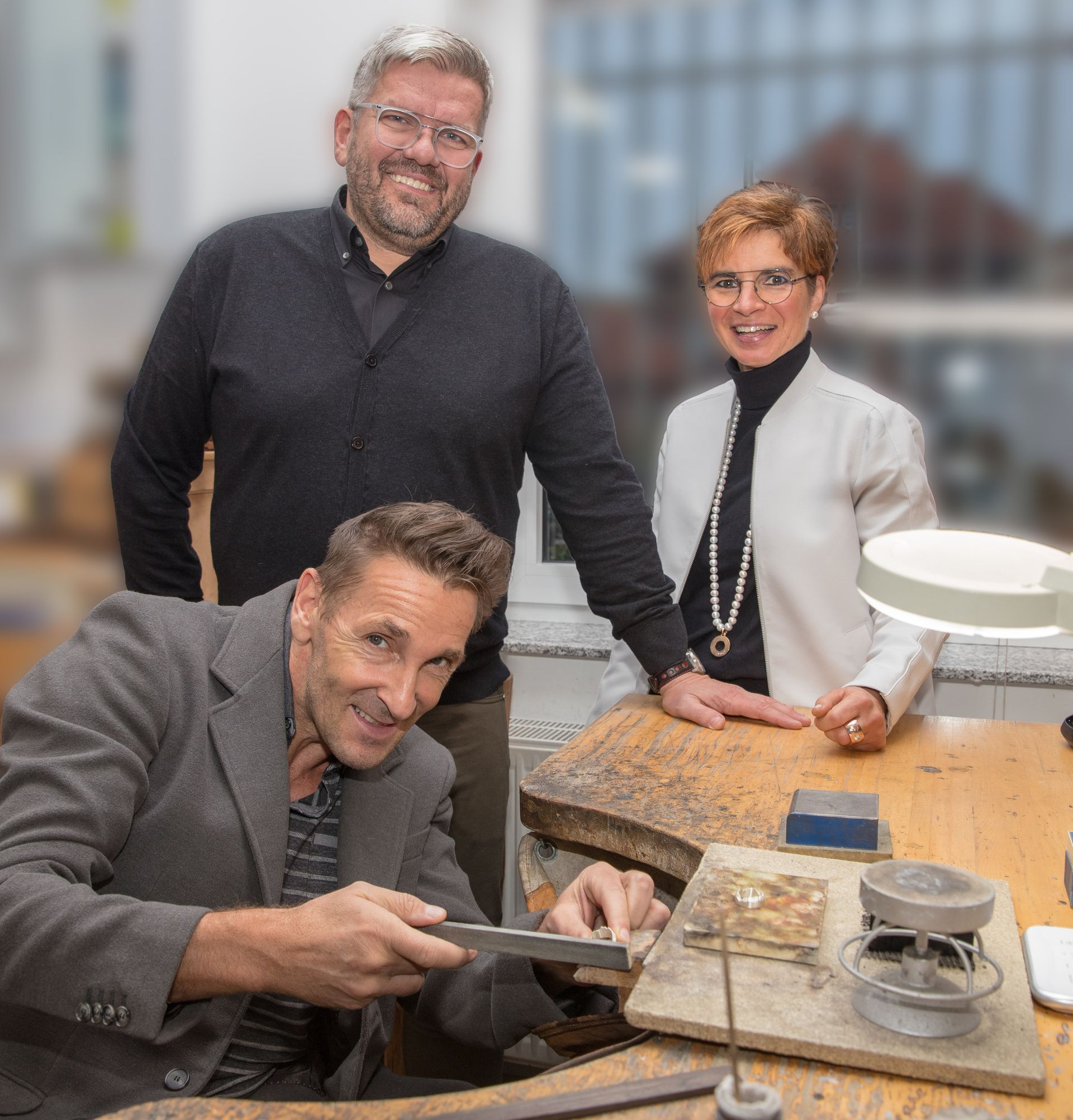 Ein Mann arbeitet an einem Stück Metall, während zwei andere Personen hinter ihm stehen