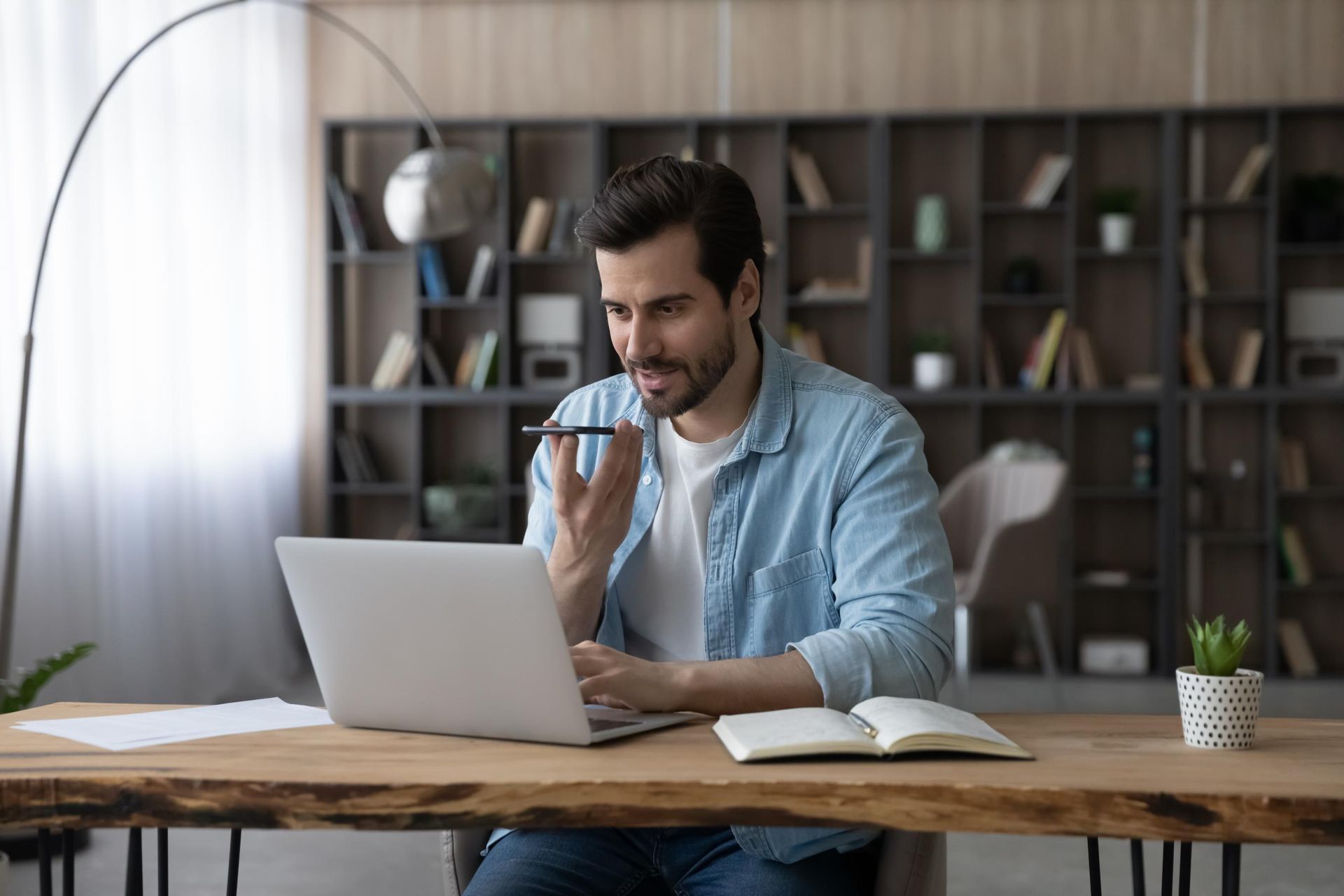 Man in blue shirt, using laptop, speaking into a phone or recorder, at wooden desk.