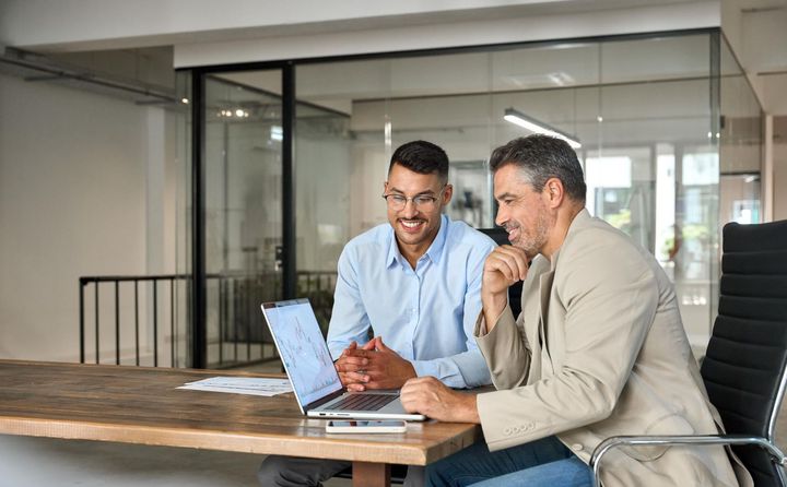 Two men at a desk with a laptop, smiling; office setting.