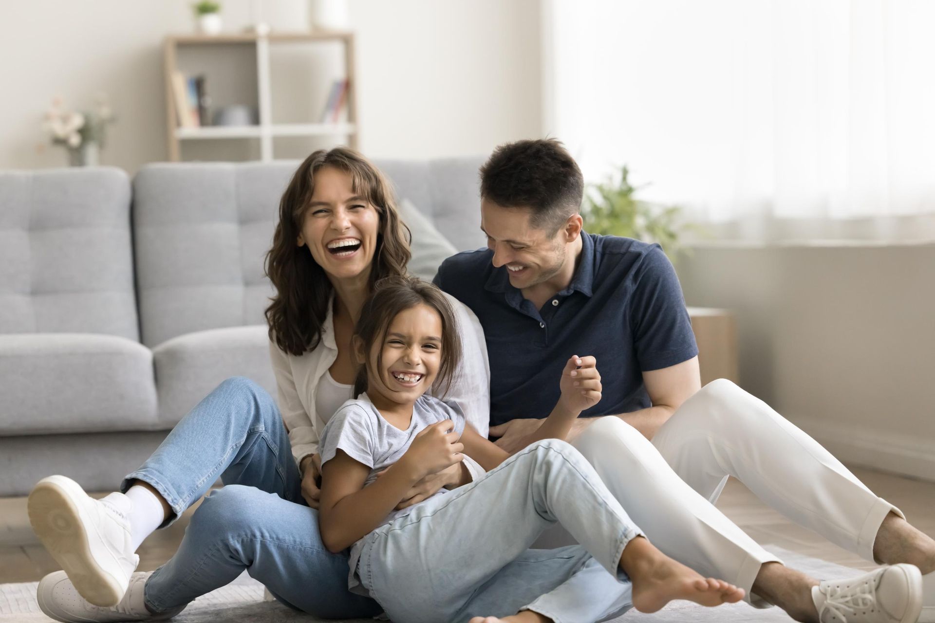 Family of three, smiling and laughing, sits on the floor in living room.